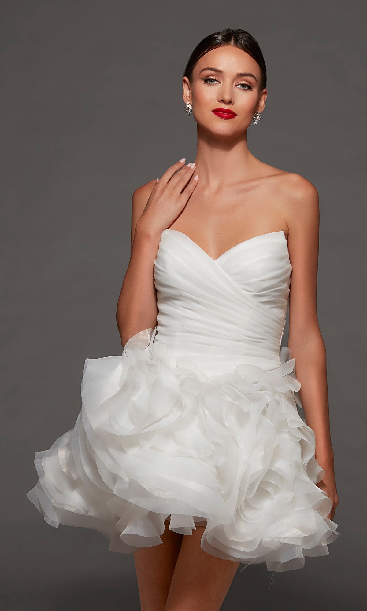 A woman wearing the Alyce 70086 short white party dress, featuring a strapless sweetheart neckline and ruffled skirt, accessorized with red lipstick and earrings, poses against a plain gray background.