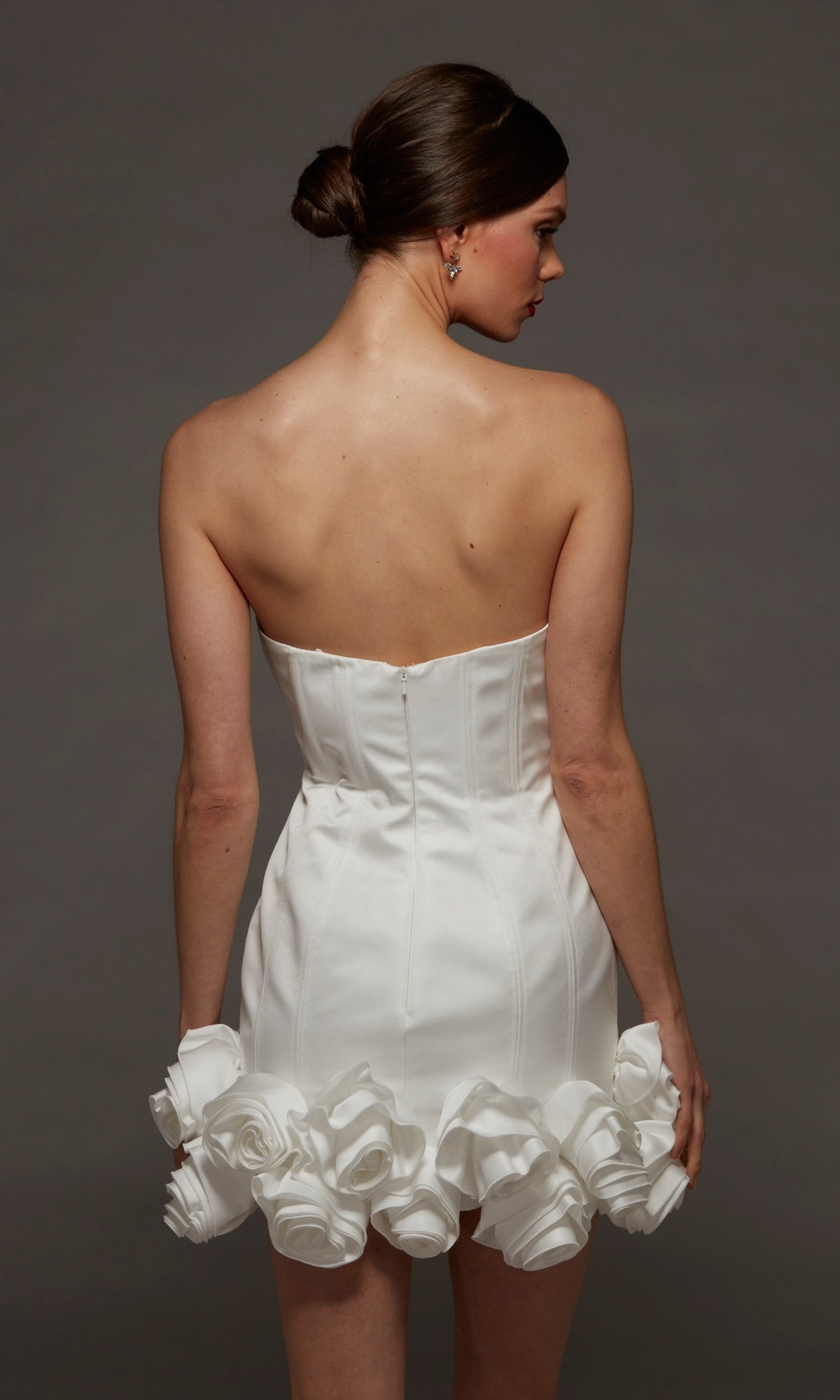 A woman faces away, wearing the Alyce 70087 short white party dress featuring a strapless bodycon silhouette with a fitted bodice and large rosette hemline details, set against a plain dark background.