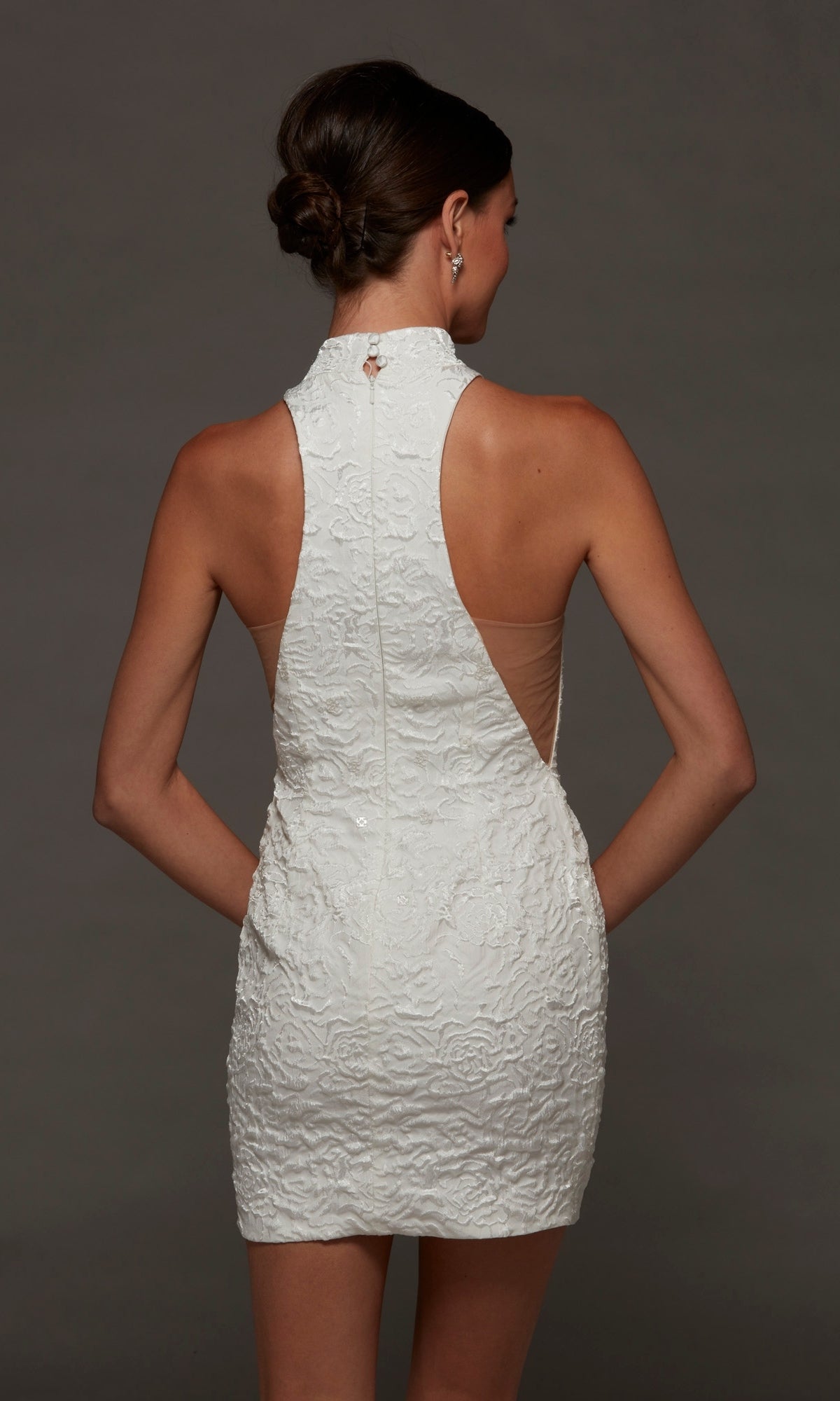 A woman stands with her back to the camera, wearing the Alyce 70094 short white party dress, a sleeveless jacquard mini with a fitted silhouette, high neckline, and nude mesh side panels.
