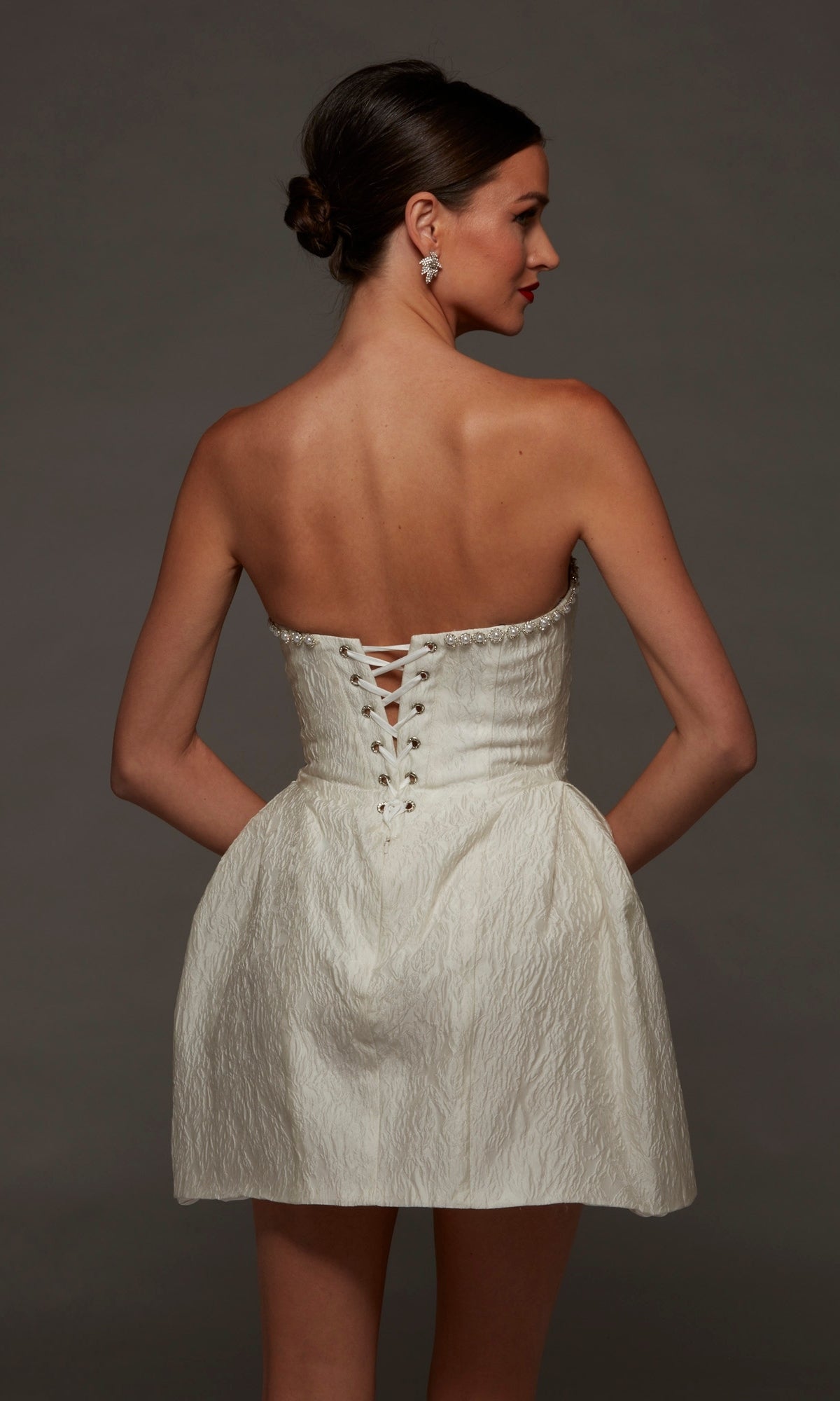 A woman is wearing the Alyce 70097 short white party dress, a strapless jacquard mini dress with a textured finish and lace-up back detail. Standing against a dark background with her hands on her hips, she showcases this perfect short party dress for any occasion.