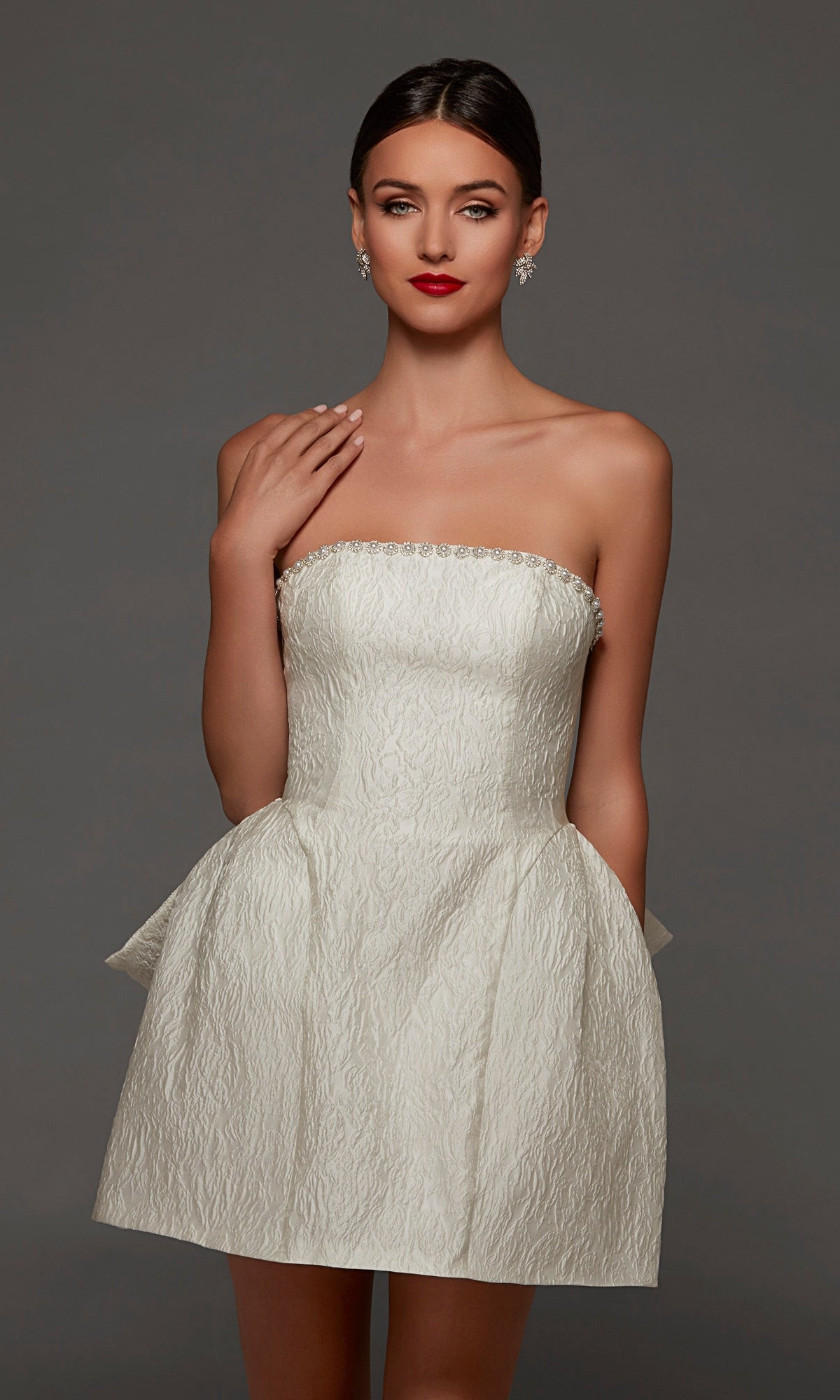 A woman wearing the Alyce 70097 Short White Party Dress, featuring a strapless, textured mini silhouette with a flared skirt and statement bow detail, stands against a gray background.
