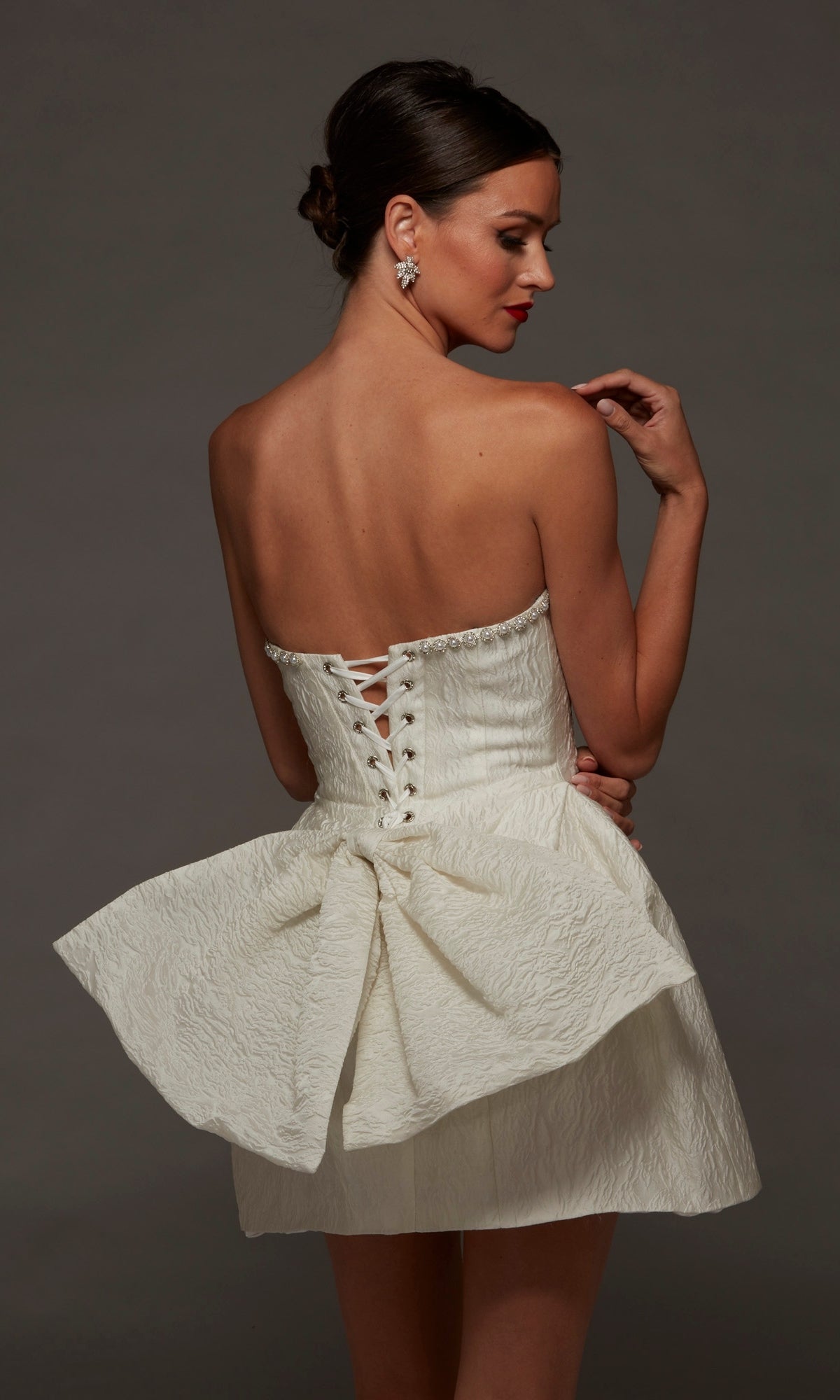 Woman wearing the Alyce 70097 short white party dress—strapless with jacquard fabric and a large bow at the back—posing against a plain dark background.