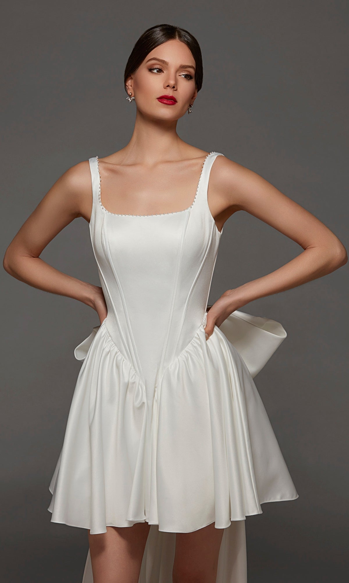 A woman models the Alyce 70103 Short White Party Dress in Diamond White, featuring a corset bodice, wide straps, and a flared skirt with a side bow detail—all elegantly framed by a chic square neckline.