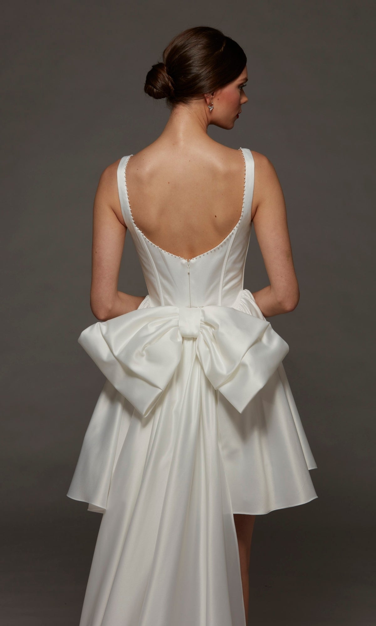 A woman wearing the Alyce 70103 Short White Party Dress, featuring a sleeveless corset bodice and a large bow at the back, stands facing away from the camera against a dark gray background.