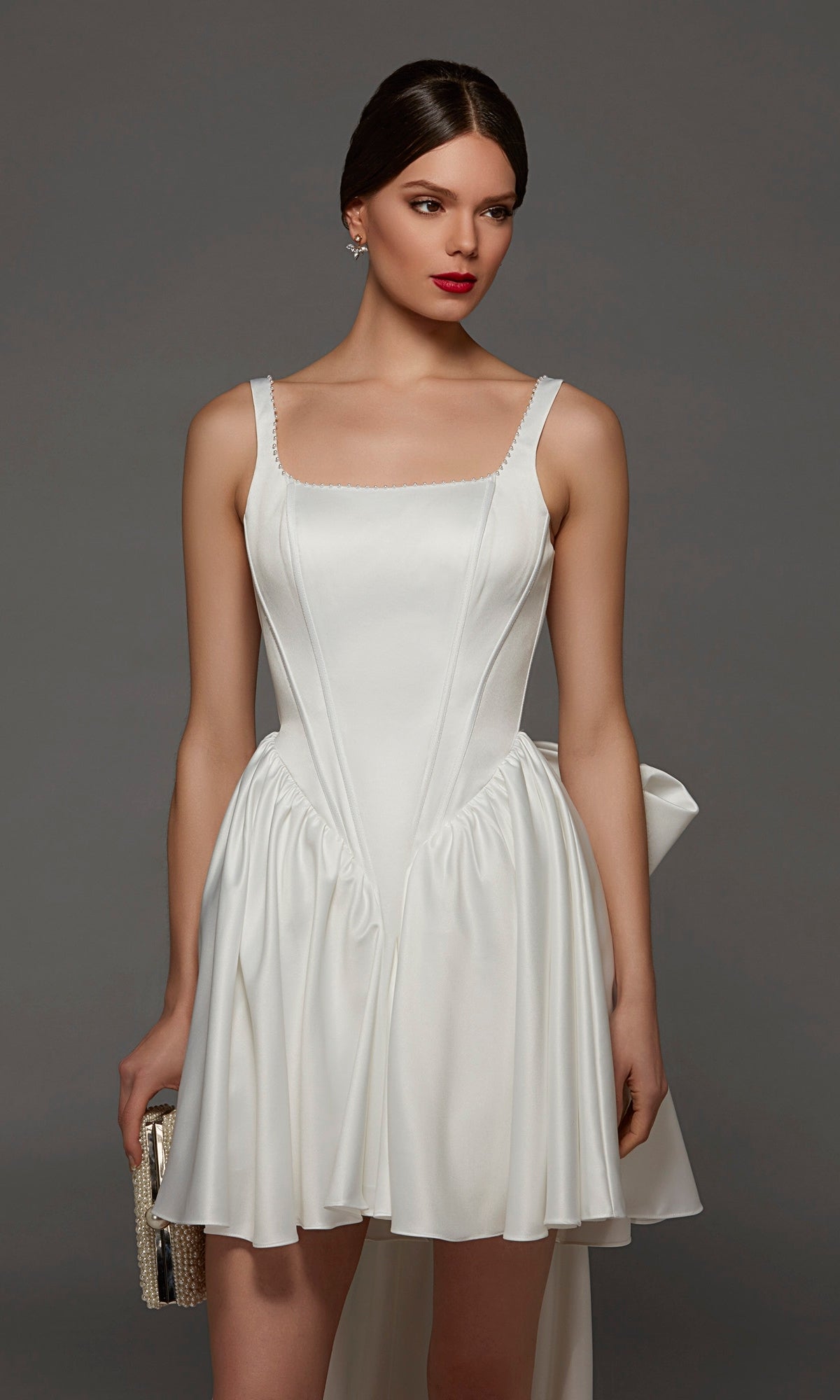 A woman wearing the Short White Party Dress: Alyce 70103—with its corset bodice, square neckline, flared skirt, and bow detail—holds a clutch against a gray background.