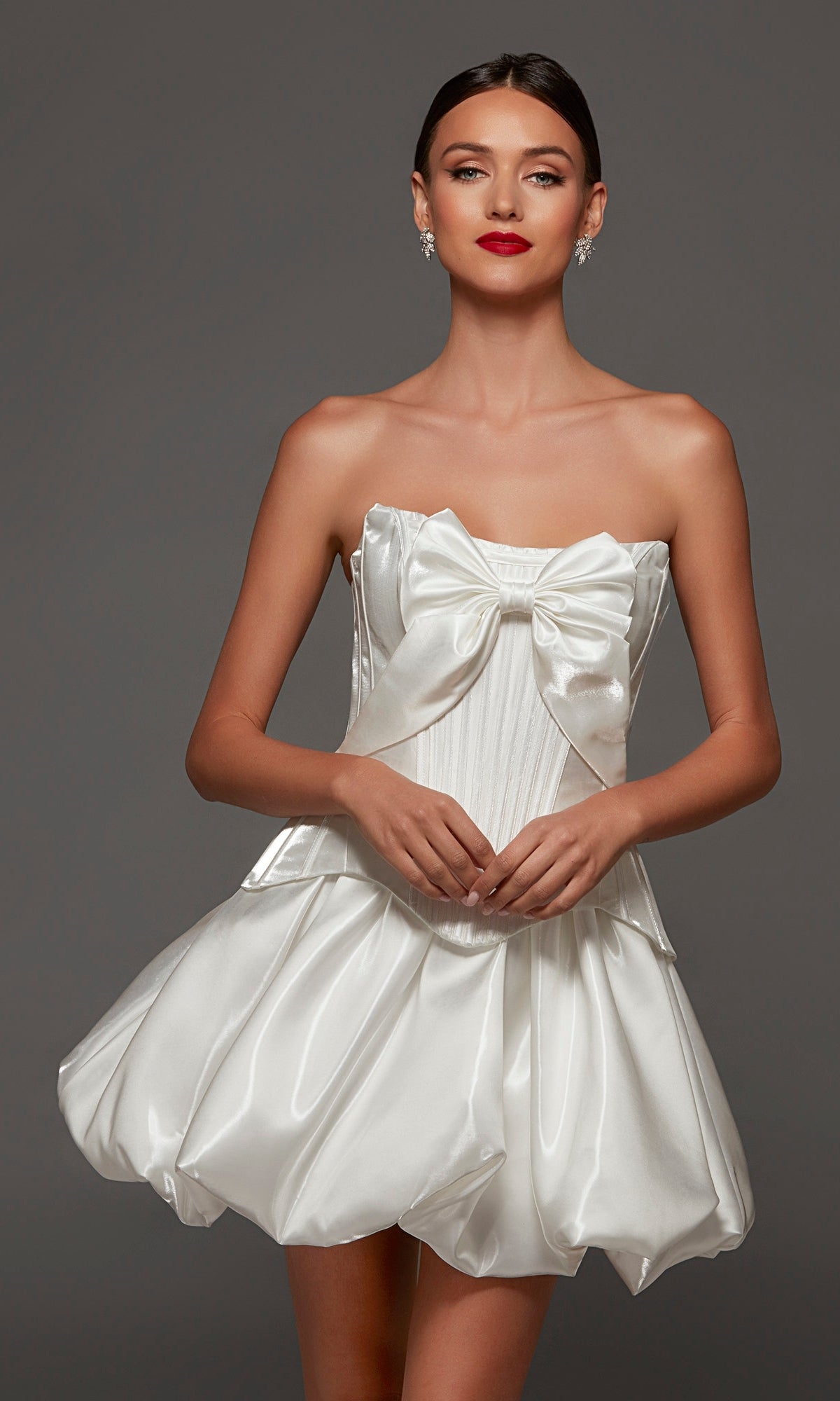 A woman wearing the Short Party Dress: Alyce 70108, a white strapless corset dress made from liquid satin with a dramatic front bow and a playful bubble-hem skirt, stands against a plain gray background.