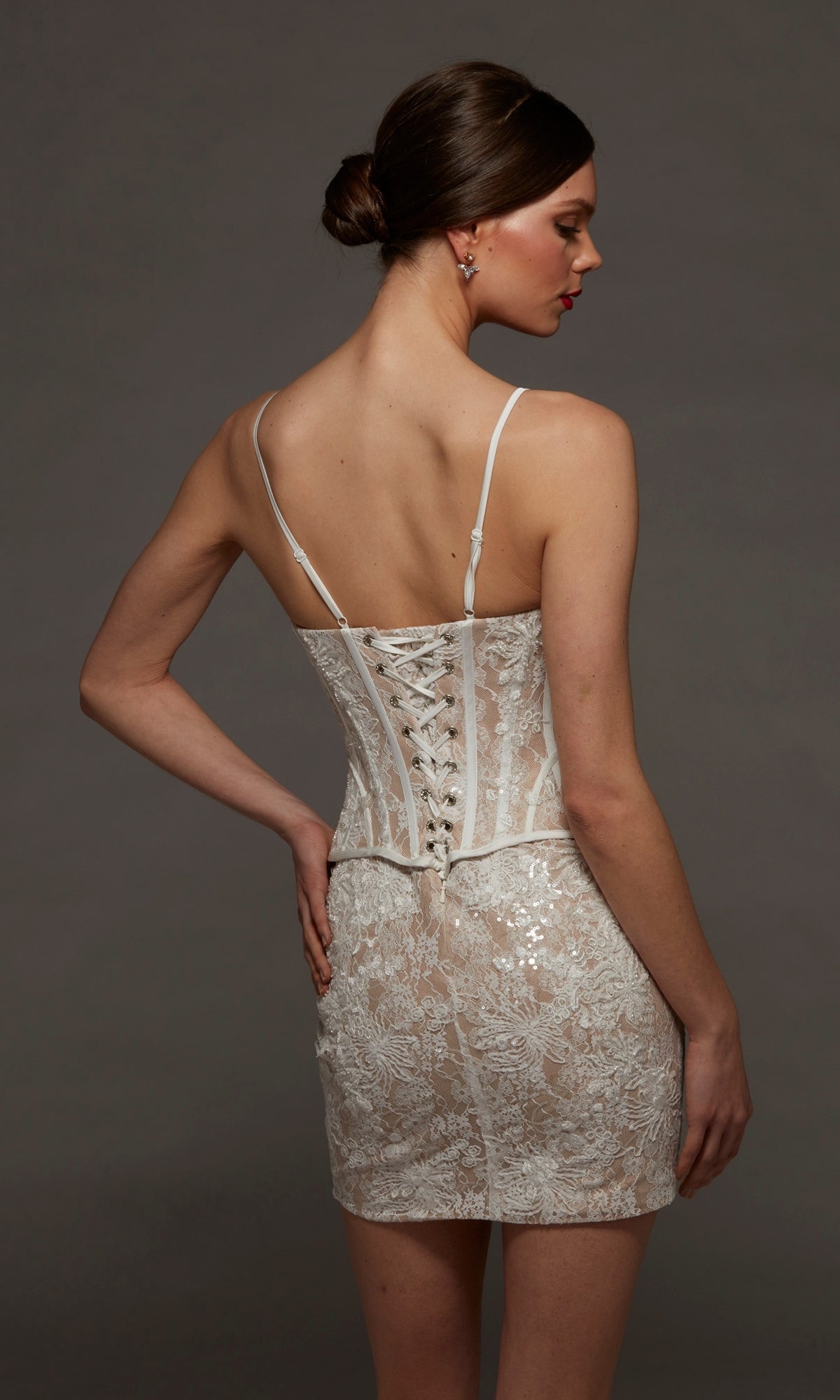 A woman stands with her back to the camera against a plain dark background, wearing the Alyce 70115 short white party dress featuring delicate lace and a lace-up back.