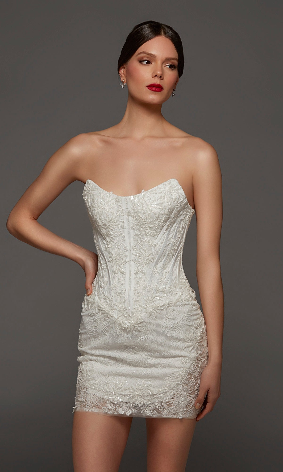 A woman stands against a plain gray background wearing the Alyce 70116 short white party dress, featuring a strapless, form-fitting silhouette with intricate lace detailing, a corset bodice, and a flattering strapless scoop neckline.
