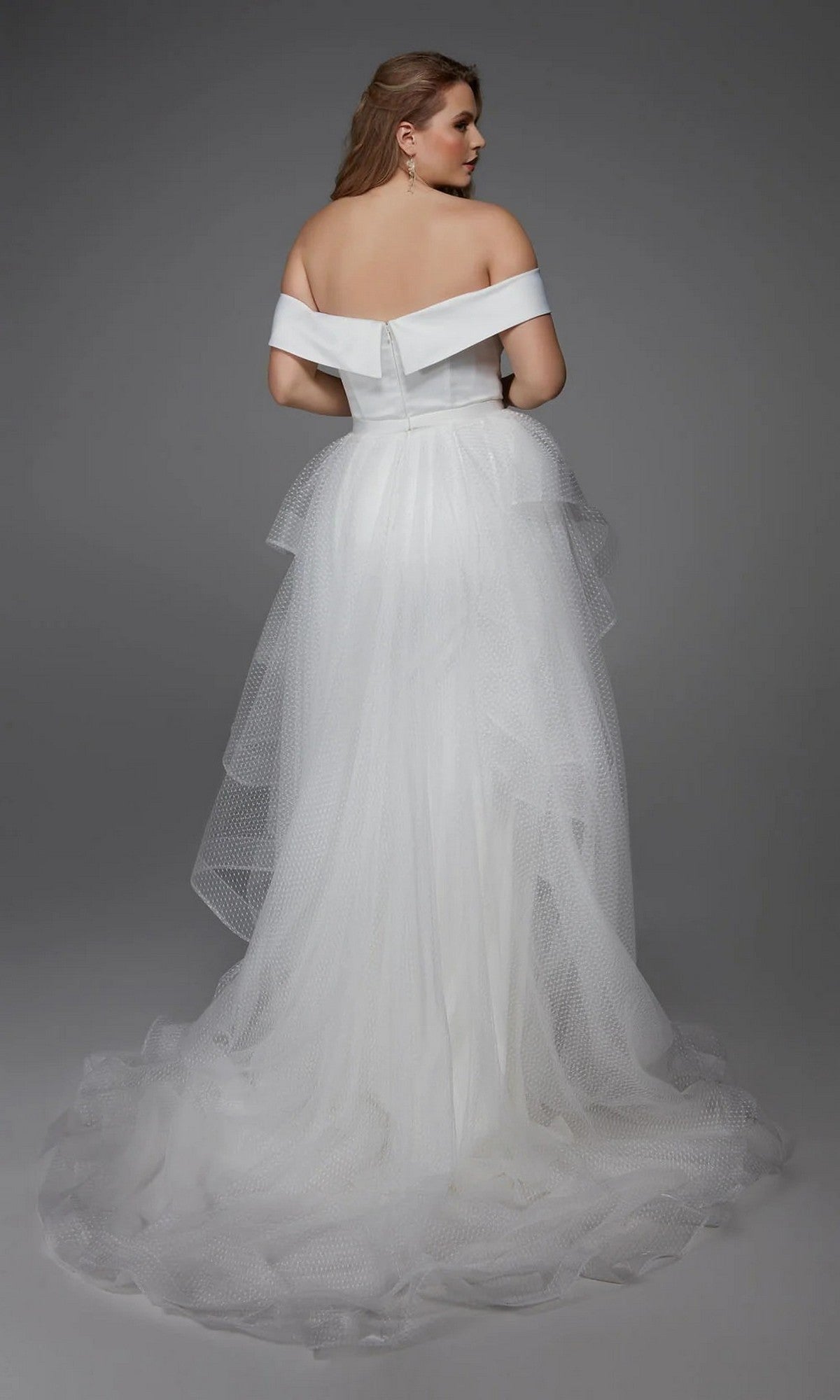 A woman models the Alyce 7020, a long white off-the-shoulder satin gown with a layered tulle skirt, standing and facing away against a gray background—ideal for wedding receptions or bridal events.