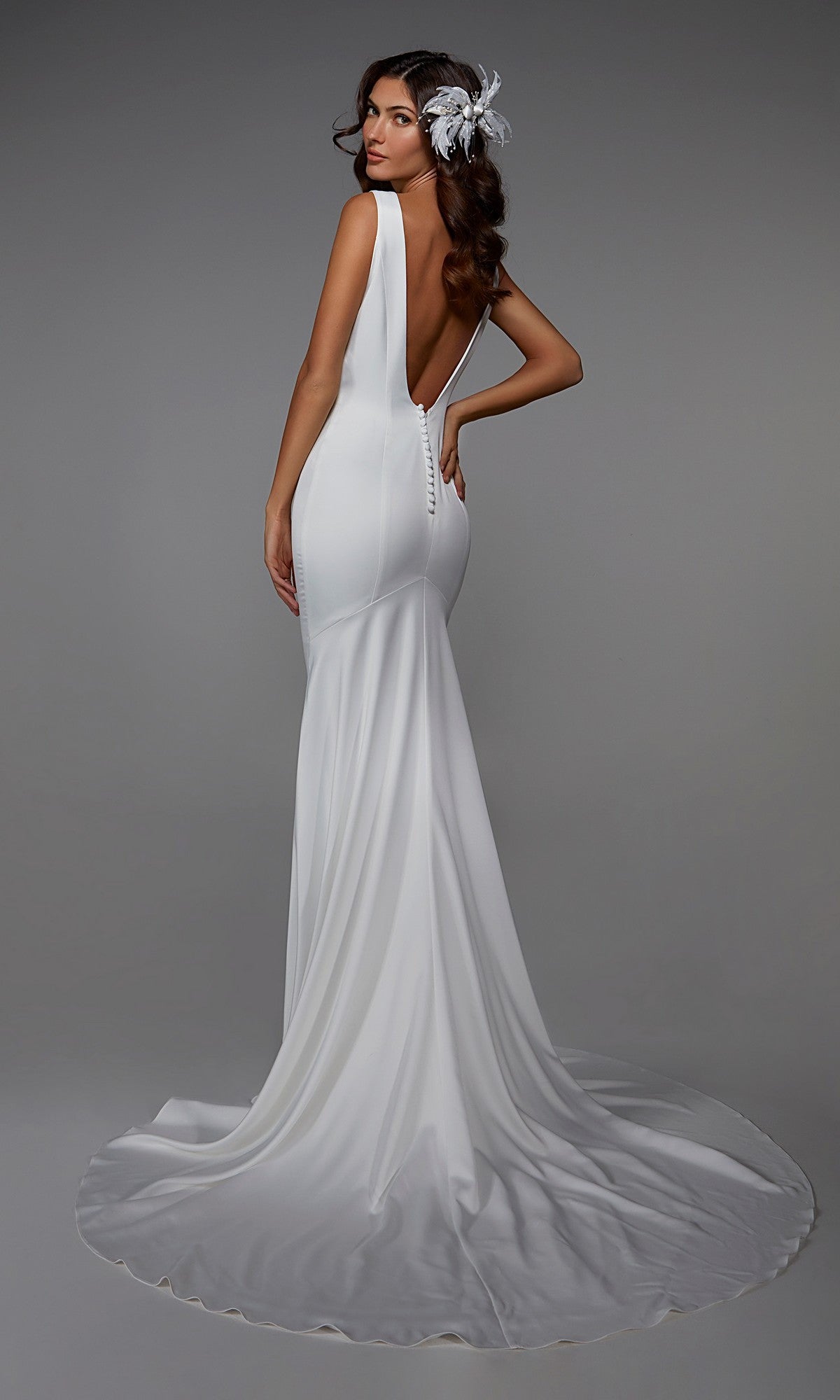 A woman stands against a plain gray background wearing the Alyce 7021 long formal dress, a white satin fit-and-flare gown with a plunging V-neckline, backless design, long train, and feather hair accessory.
