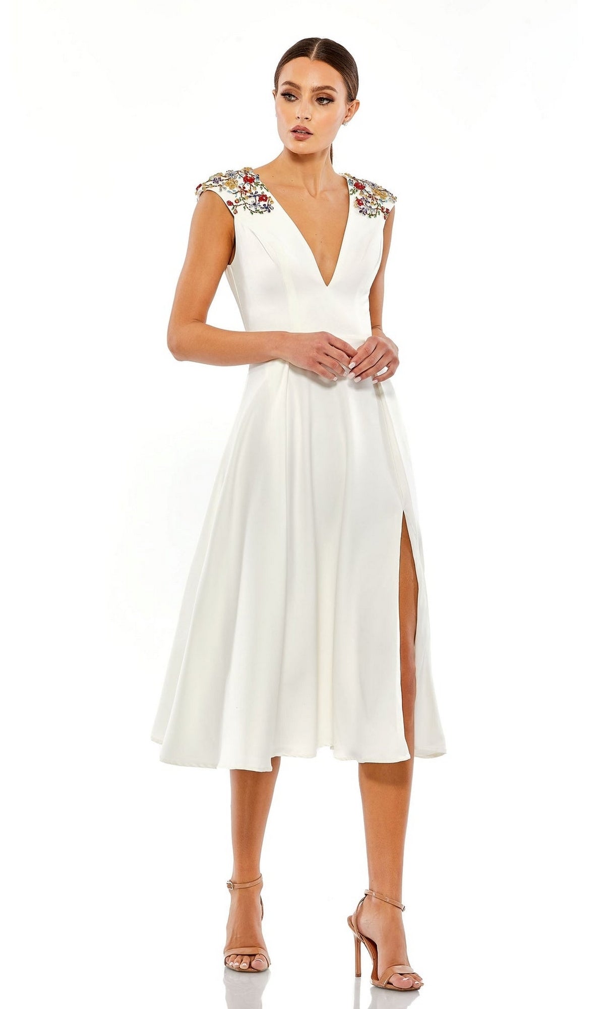 A woman wears the Short Wedding Guest Dress 70214 by Mac Duggal, a white beaded knee-length party dress with a deep V-neck, floral shoulder embellishments, front slit, and nude high heels.