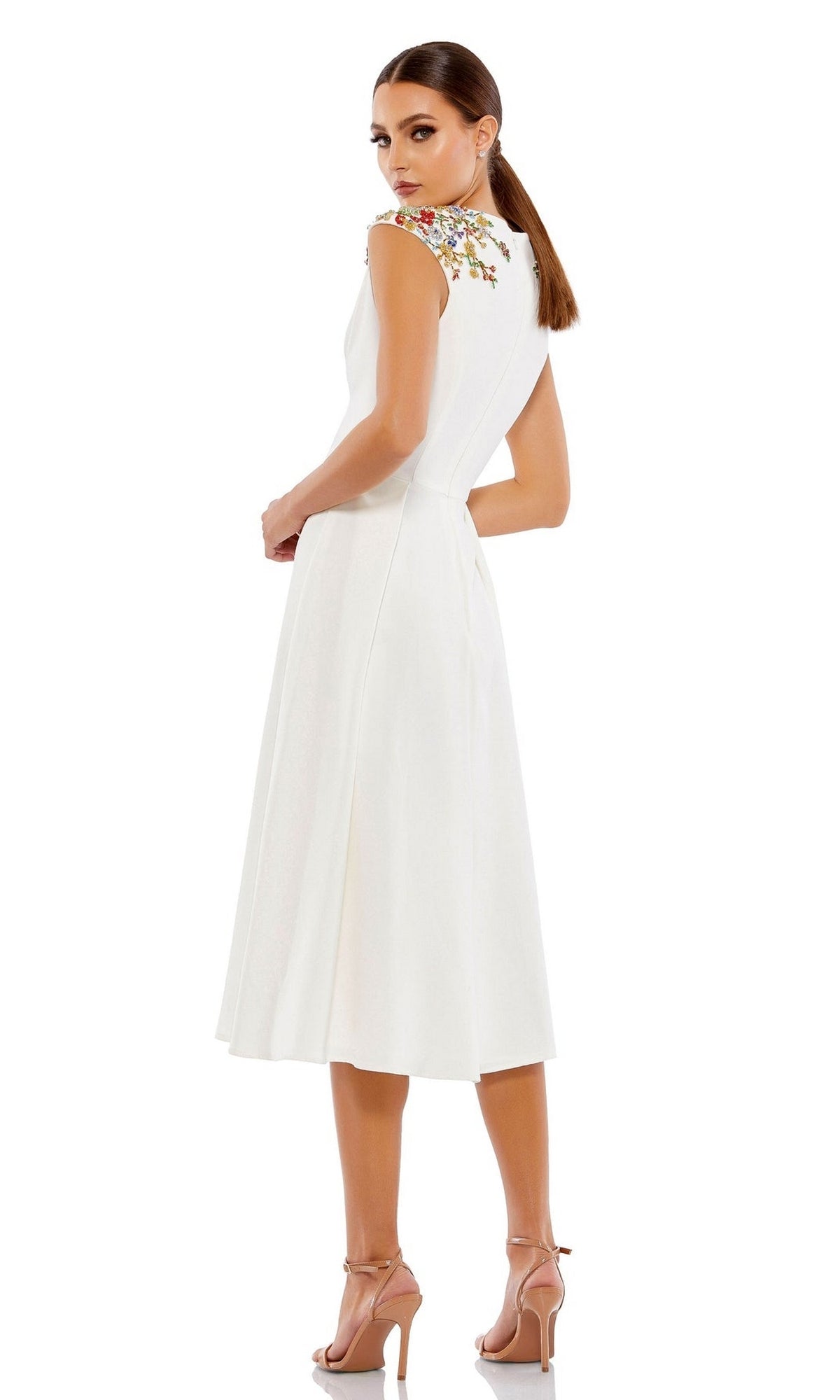 A woman stands sideways and looks back in the Short Wedding Guest Dress 70214 by Mac Duggal, a white sleeveless knee-length v-neck A-line dress with floral embroidery on the shoulders, paired with nude high heels against a plain background.