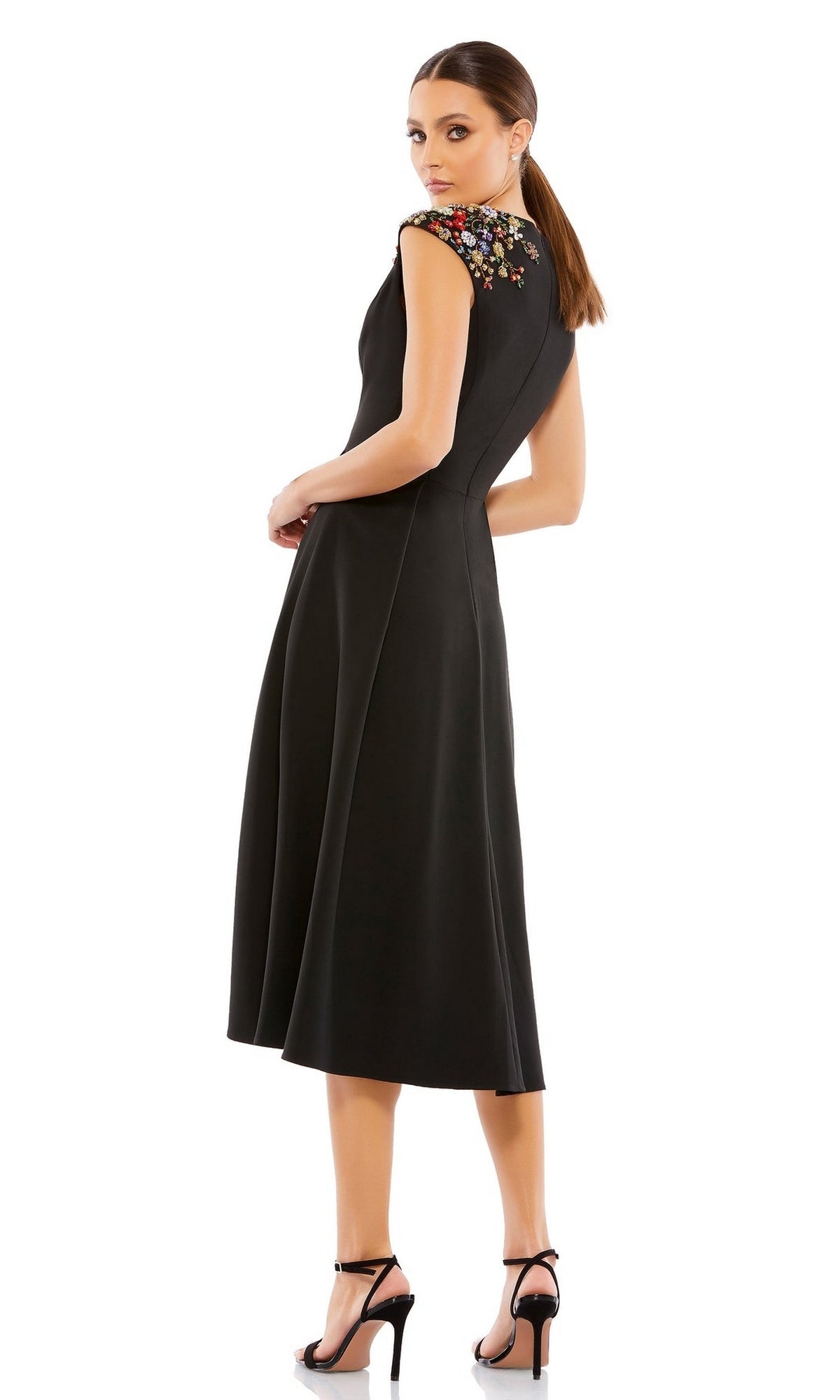 A woman models the Mac Duggal Short Wedding Guest Dress 70214—a black, knee-length v-neck A-line dress with colorful floral shoulder embroidery—standing in heels and looking over her shoulder against a white background.