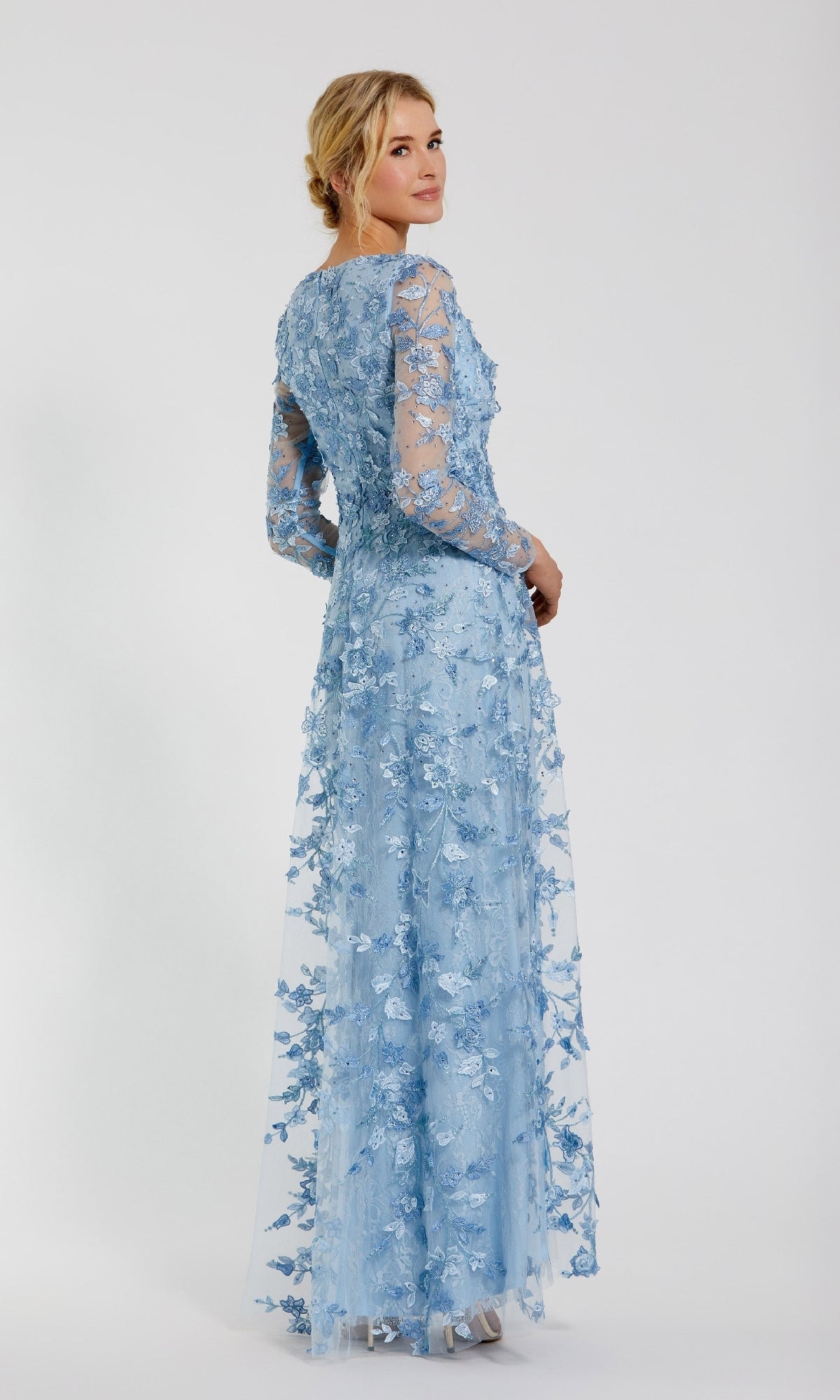 A woman stands slightly turned away, wearing the Mac Duggal 70224 Long Sleeve Long Formal Dress in light blue with floral embroidery and sheer sleeves, set against a plain white background.
