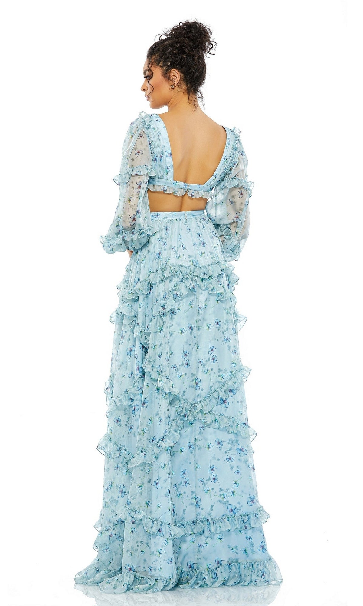 A woman faces away, wearing the Mac Duggal 70249 long sleeve blue formal dress featuring a floral print, ruffles, puffed sleeves, and an open back finished with a bow.