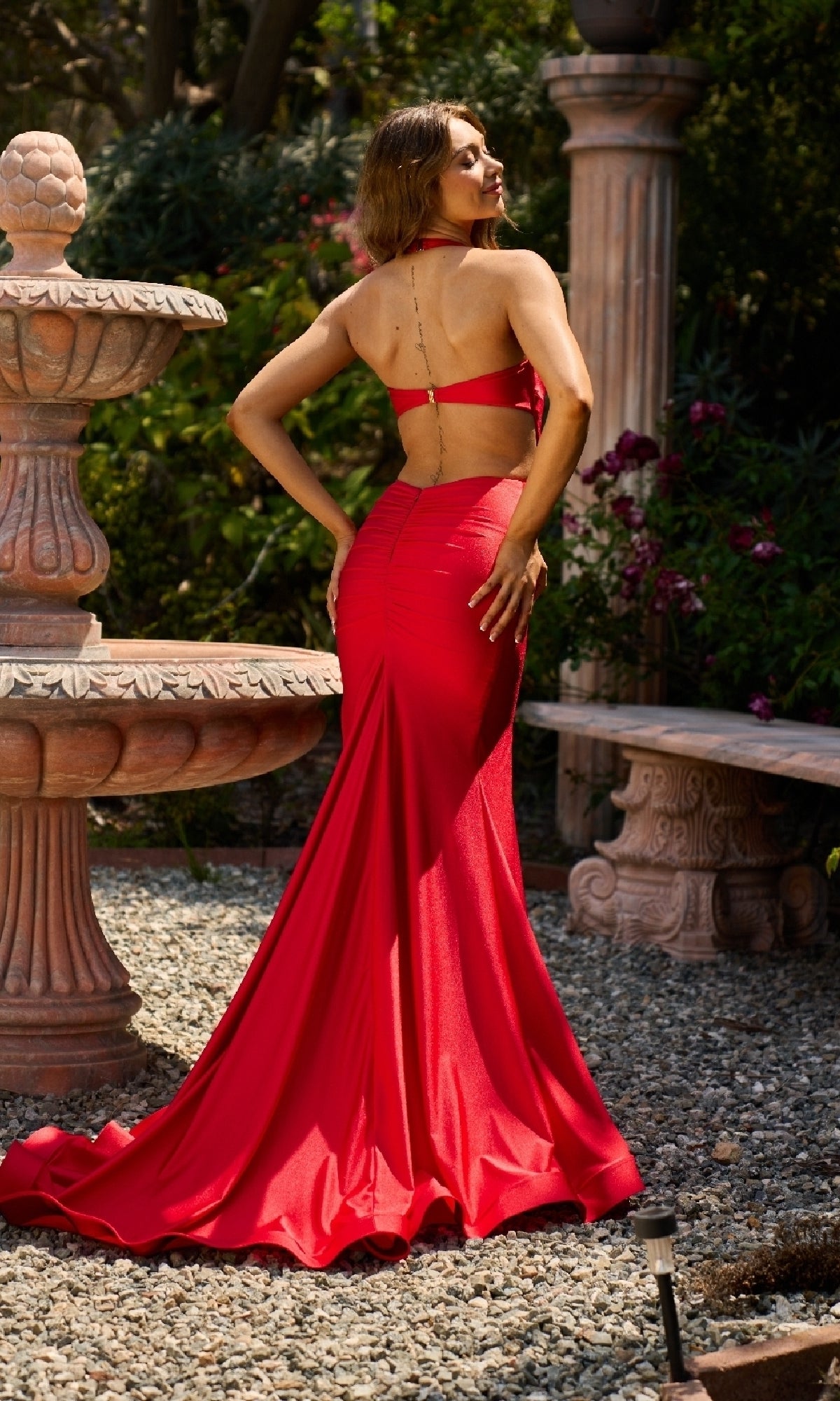 A woman wearing the Atria 7034H Long Halter Prom Dress stands in a garden near a stone fountain and bench, facing away from the camera.