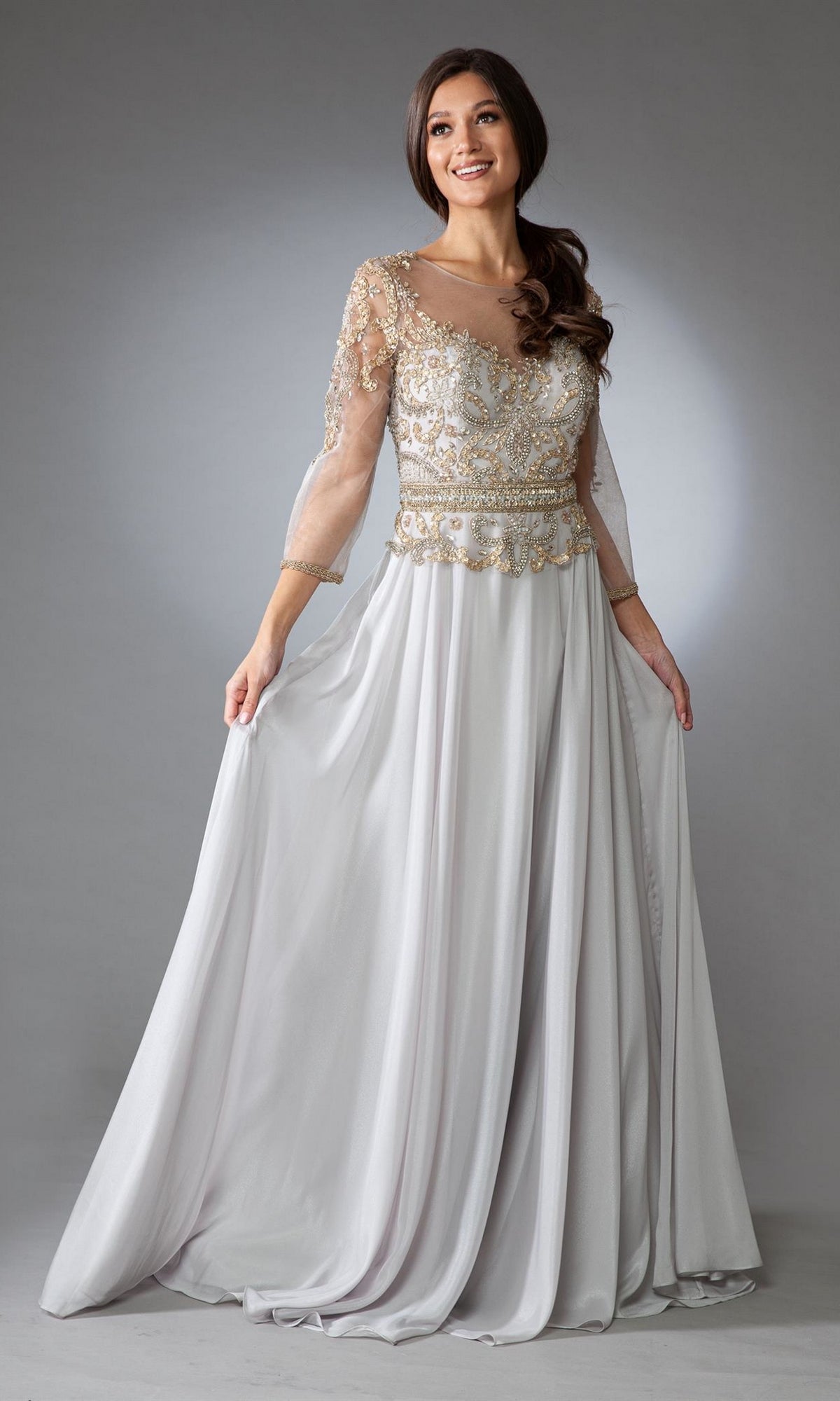 A woman wears the Sheer-Sleeve Long Modest Formal Dress 7041, a light gray gown featuring flowing fabric, sheer embroidered sleeves, and intricate gold detailing on the bodice, standing against a plain background.