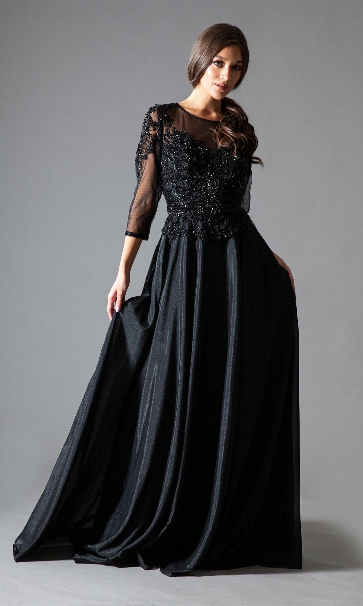 A woman models the Sheer-Sleeve Long Modest Formal Dress 7041, featuring sheer sleeves and an intricate beaded bodice, against a plain gray background.