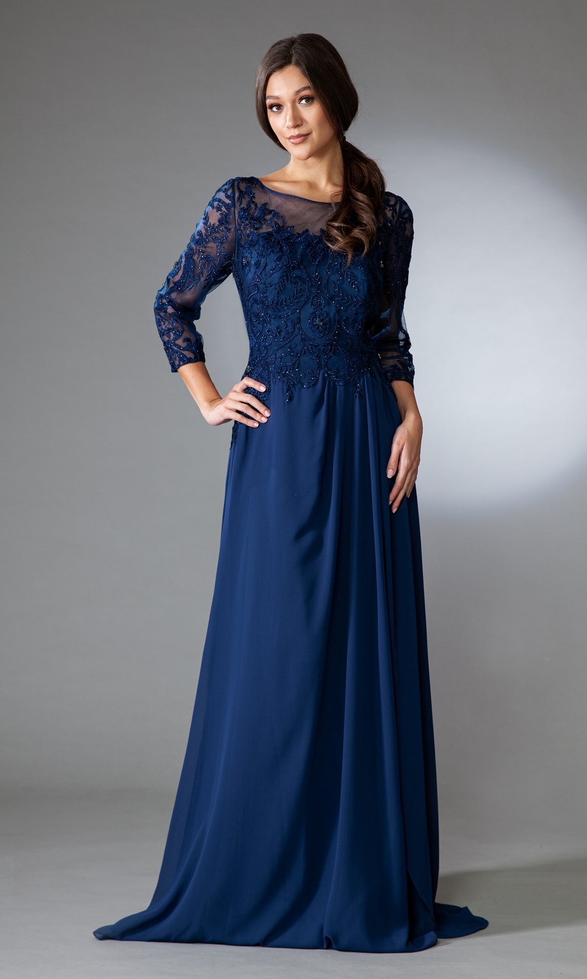 A woman poses against a neutral background in the affordable Modest Long-Sleeve Mother-of-the-Bride Dress 7043—a floor-length navy blue gown with sheer lace sleeves and intricate bodice embroidery.