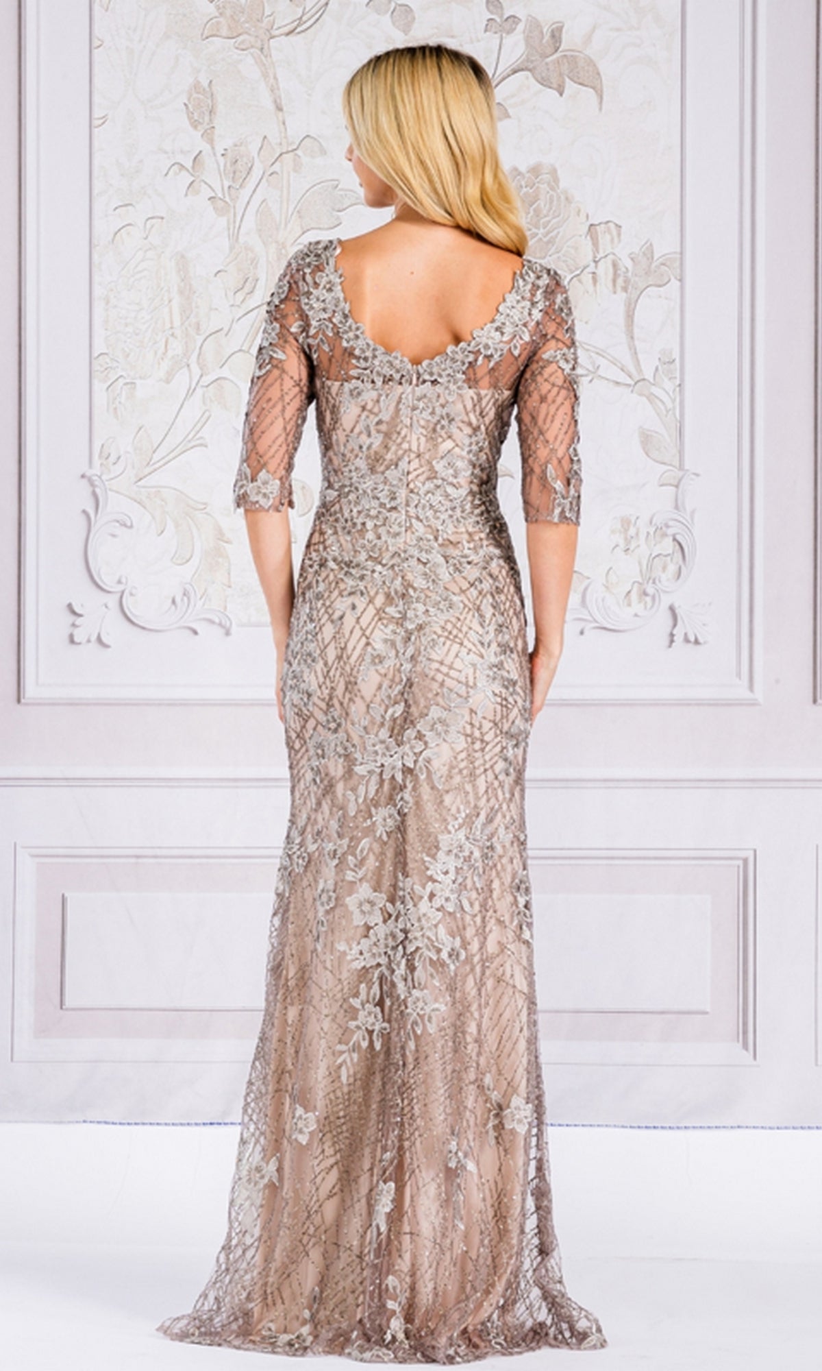 A woman stands with her back to the camera wearing the Floral-Embroidered Long Sleeve Formal Dress 7045, a floor-length light brown gown featuring silver floral embroidery and sheer sleeves.