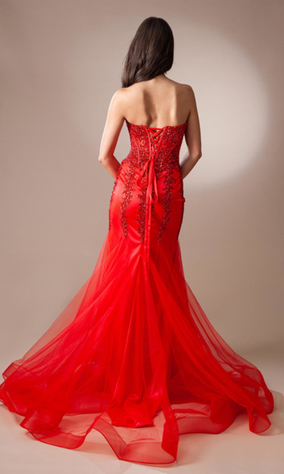 A woman with long brown hair is seen from the back wearing the Strapless Long Beaded Sheer-Bodice Prom Dress 7051, which features a form-fitting red design with a corset-style bodice, black lace accents, and a tulle train.