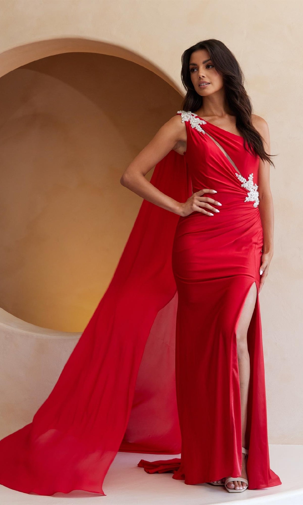 A woman in the Amelia Couture 7056, a bright red one-shoulder long prom dress with white floral details, flowing cape, and thigh-high slit, stands indoors against a curved beige backdrop.