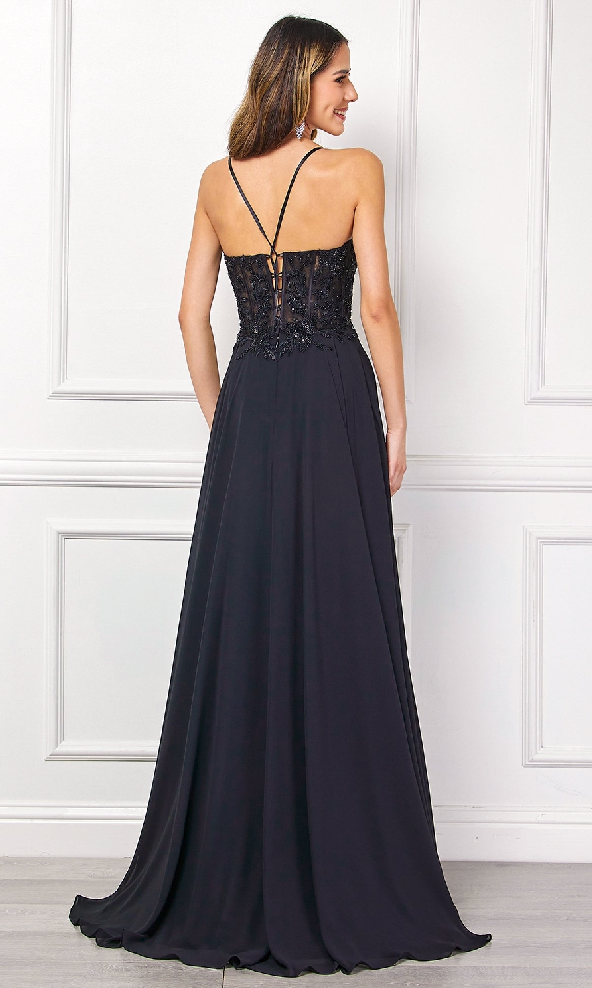 A woman faces away, wearing the Long Black Corset Prom Dress: Amelia Couture 7066, featuring a fitted beaded corset bodice with thin straps, a flowing skirt, and open back, set against a white paneled wall.