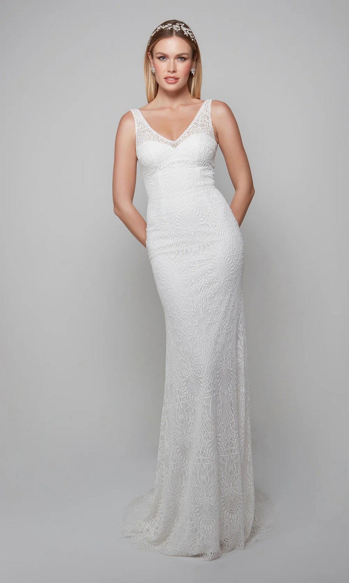 A woman in the Long White Dress: Alyce 7075, a sleeveless sheath wedding gown with lace appliqués and a V-neckline, stands against a plain light gray background.