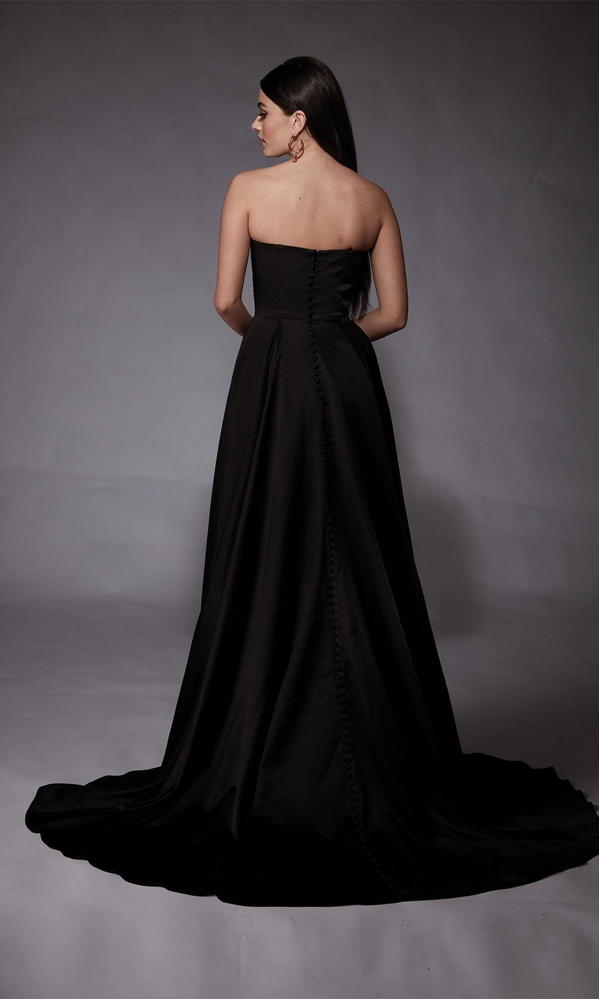 A woman with long dark hair models the Alyce 7088 long formal dress, a strapless gown with a floor-length train and button-back details, standing against a plain gray background.