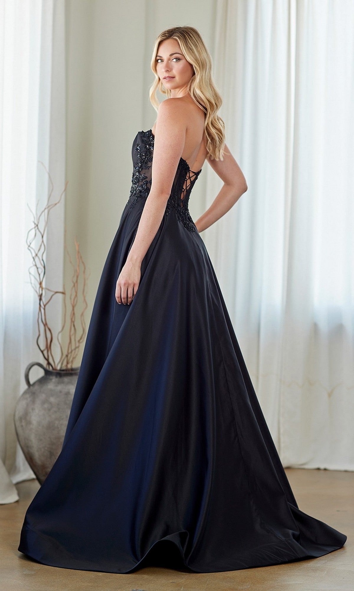 A woman models the Amelia Couture 7093, a strapless long prom dress with an embroidered corset bodice and flowing A-line skirt, in a sunlit room with white curtains and a large vase in the background.