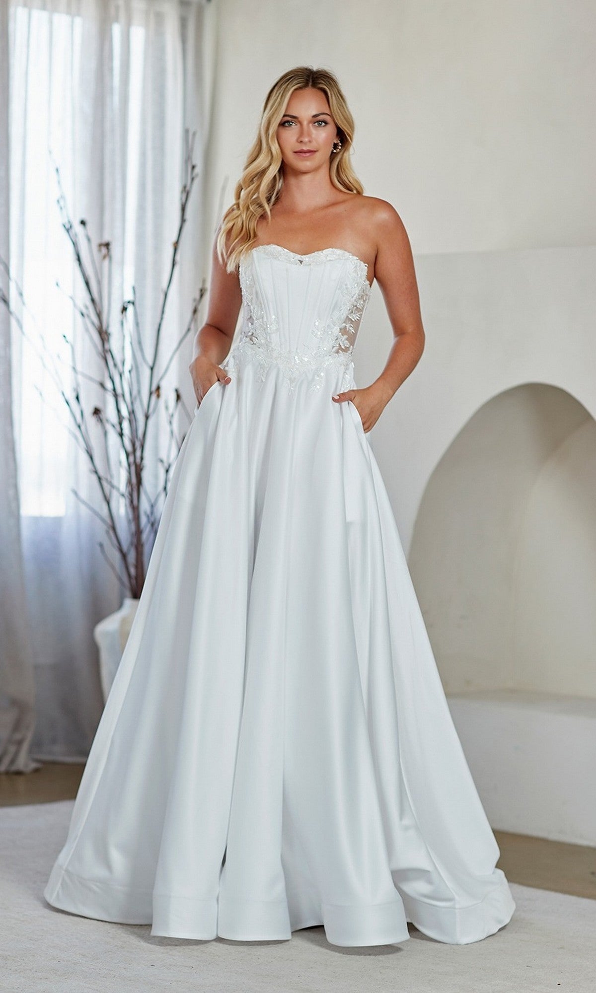 A woman stands indoors wearing the white Amelia Couture 7093 long prom dress, which features a strapless embroidered corset bodice, full A-line skirt, and pockets, set against a neutral background with curtains and decorative branches.