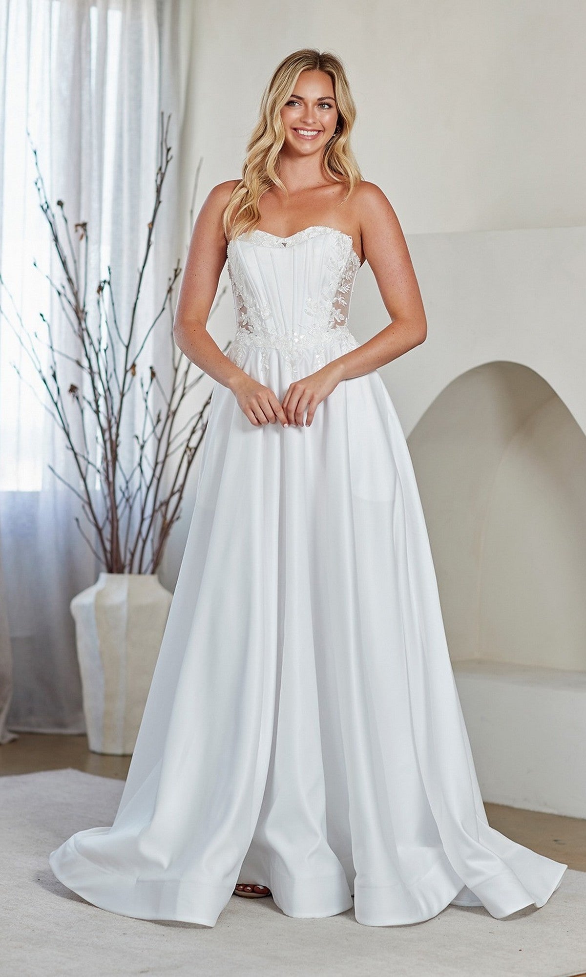 A woman stands indoors wearing the Amelia Couture 7093 long prom dress, featuring a strapless white design with an embroidered corset bodice and flowing skirt.