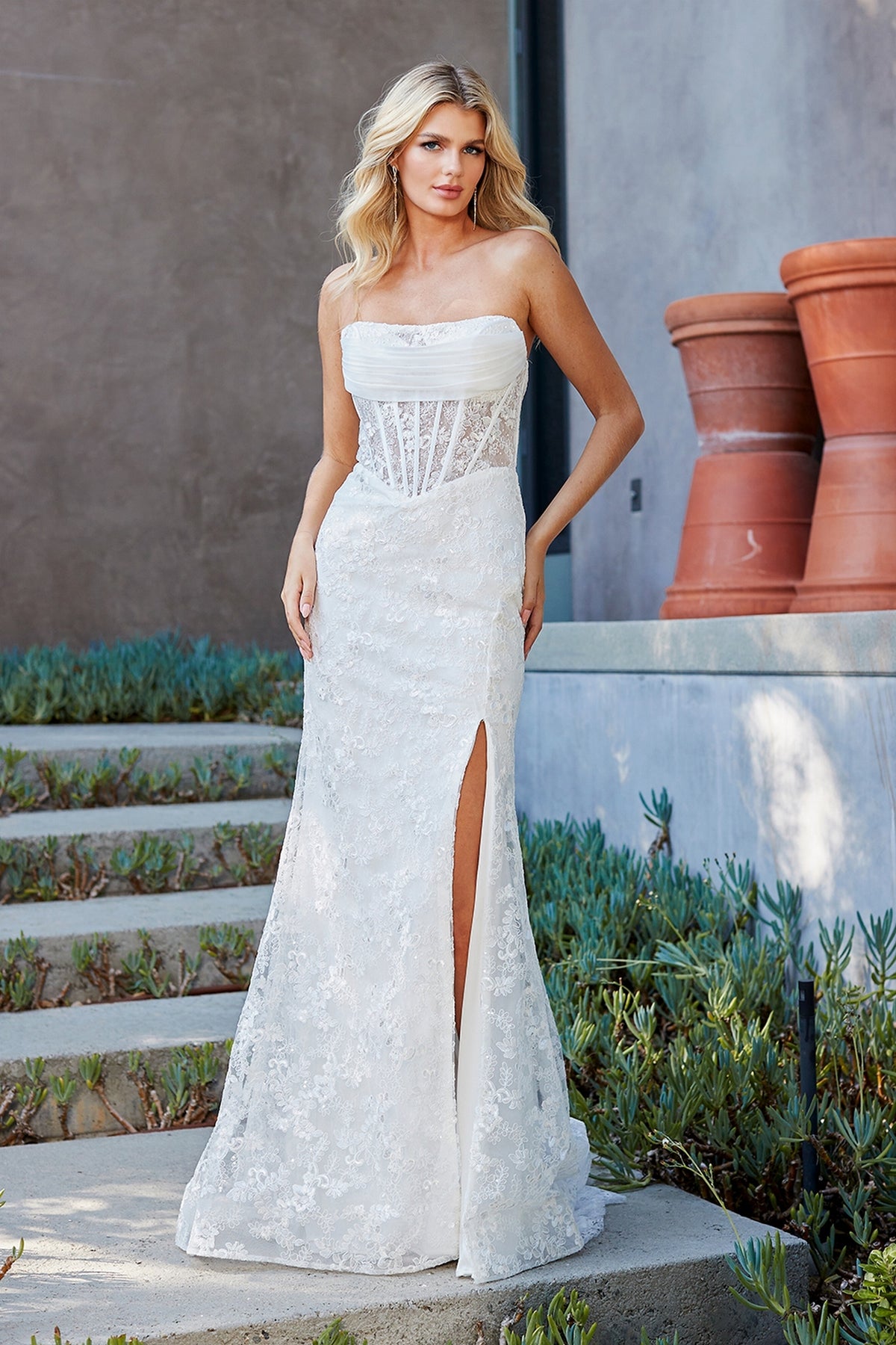 A woman wears a white Amelia Couture 7097 strapless, floor-length lace gown with a corset bodice and a high side slit.