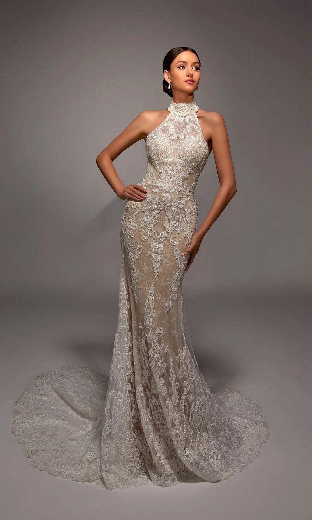 A woman models the Alyce 7159 long white dress in Ivory/Latte, featuring elegant lace, a halter neckline, and a flowing train, standing in a studio with a plain background.