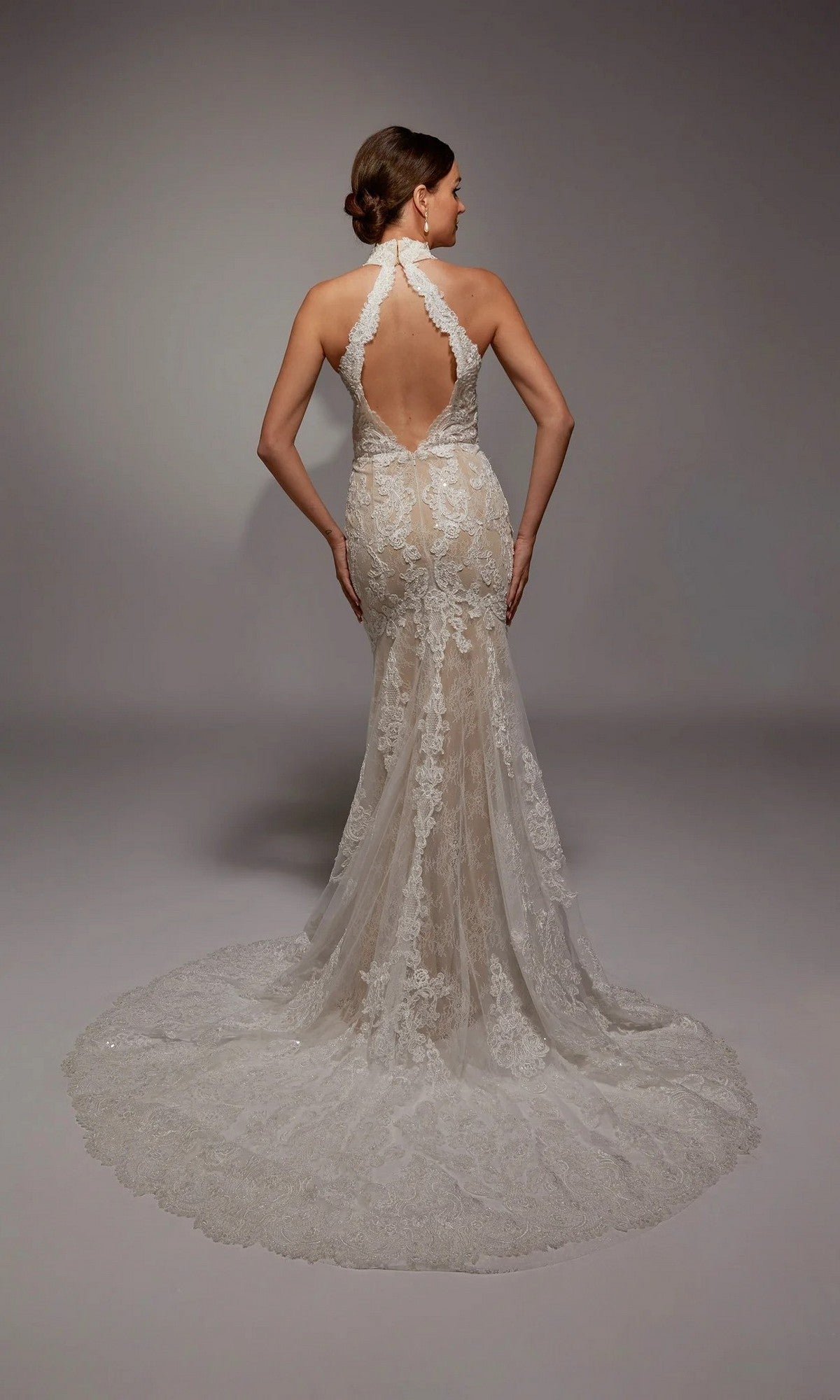 A woman wears the Alyce 7159 long white dress, featuring an ivory mermaid silhouette, lace halter-neck, open back, and long scalloped train, standing against a plain gray background.