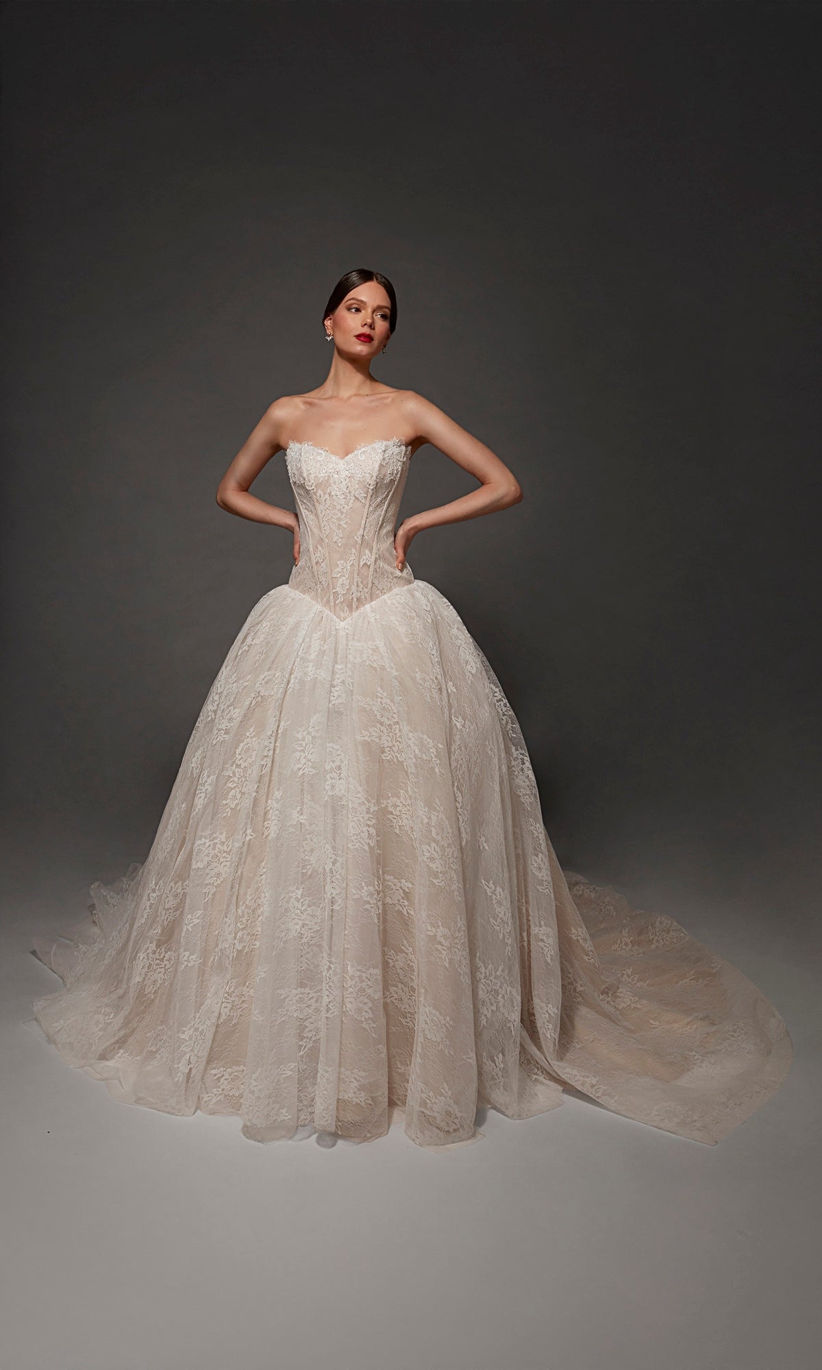 A woman wearing the Long Wedding Dress: Alyce 7169—a strapless, lace ball gown with a sweetheart neckline—stands with her hands on her hips against a plain dark background.