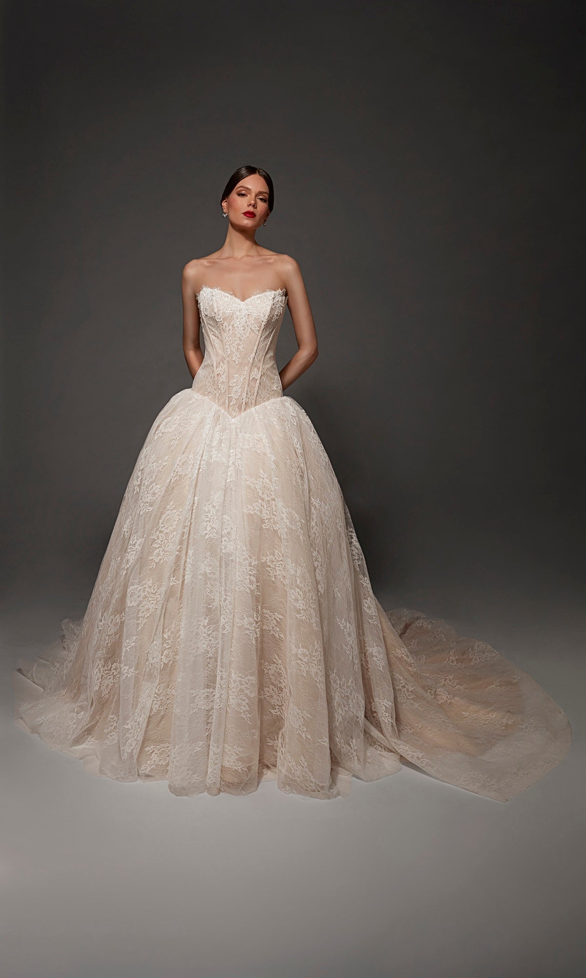 A woman stands in the Alyce 7169 long wedding dress, a strapless lace and tulle ball gown featuring a full skirt and a sweeping train, set against a dark, neutral background.