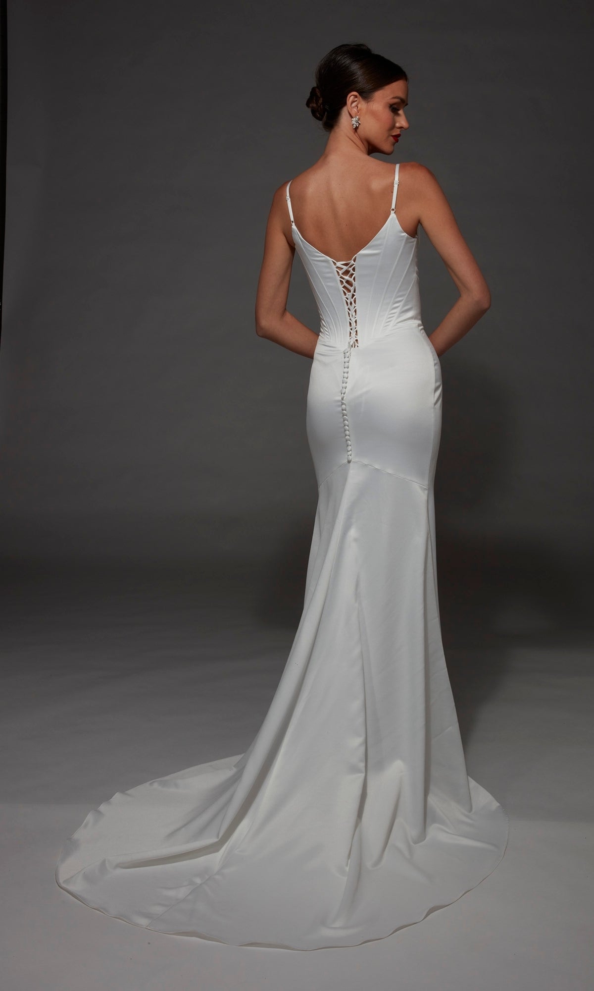 A woman in the Long Wedding Dress: Alyce 7175—a white, fitted, floor-length satin gown featuring thin straps, a graceful cowl neckline, lace-up back, and a slight train—stands against a gray background.