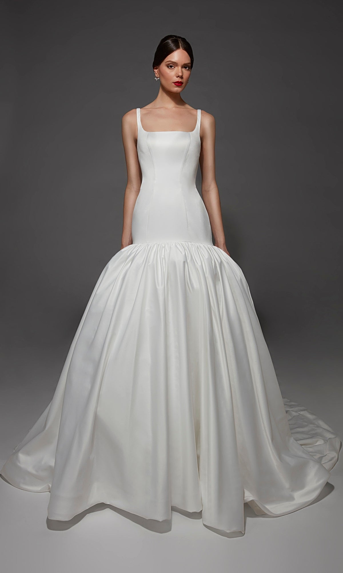 A woman wearing the Alyce 7176 long wedding dress in diamond white, a fitted, sleeveless mermaid gown with a square neckline, dramatic flared skirt, and striking train, stands against a plain gray background.