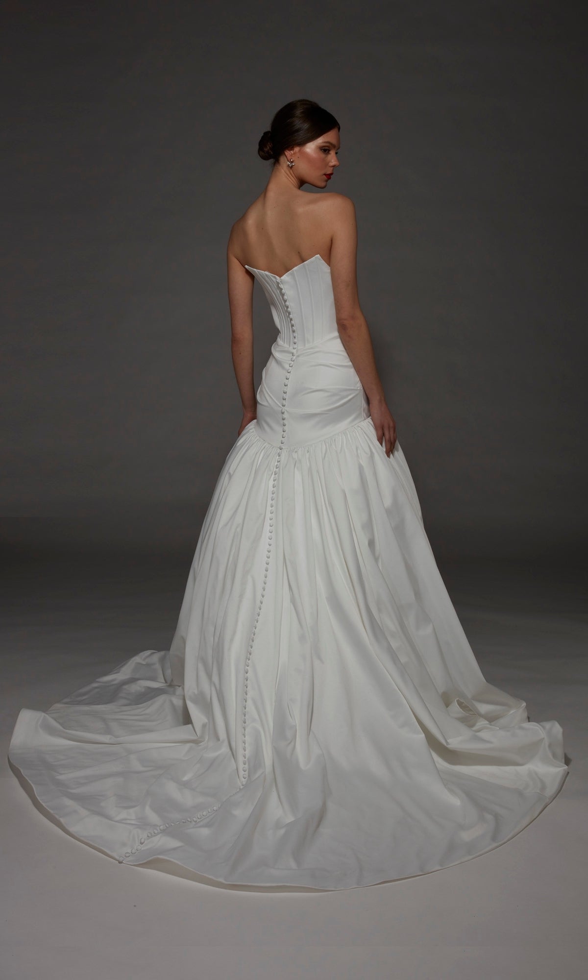 A woman stands with her back to the camera in the Alyce 7181 Long Wedding Dress, featuring a strapless satin design with a fitted bodice accented by buttons and a long, flowing train, set against a dark background.