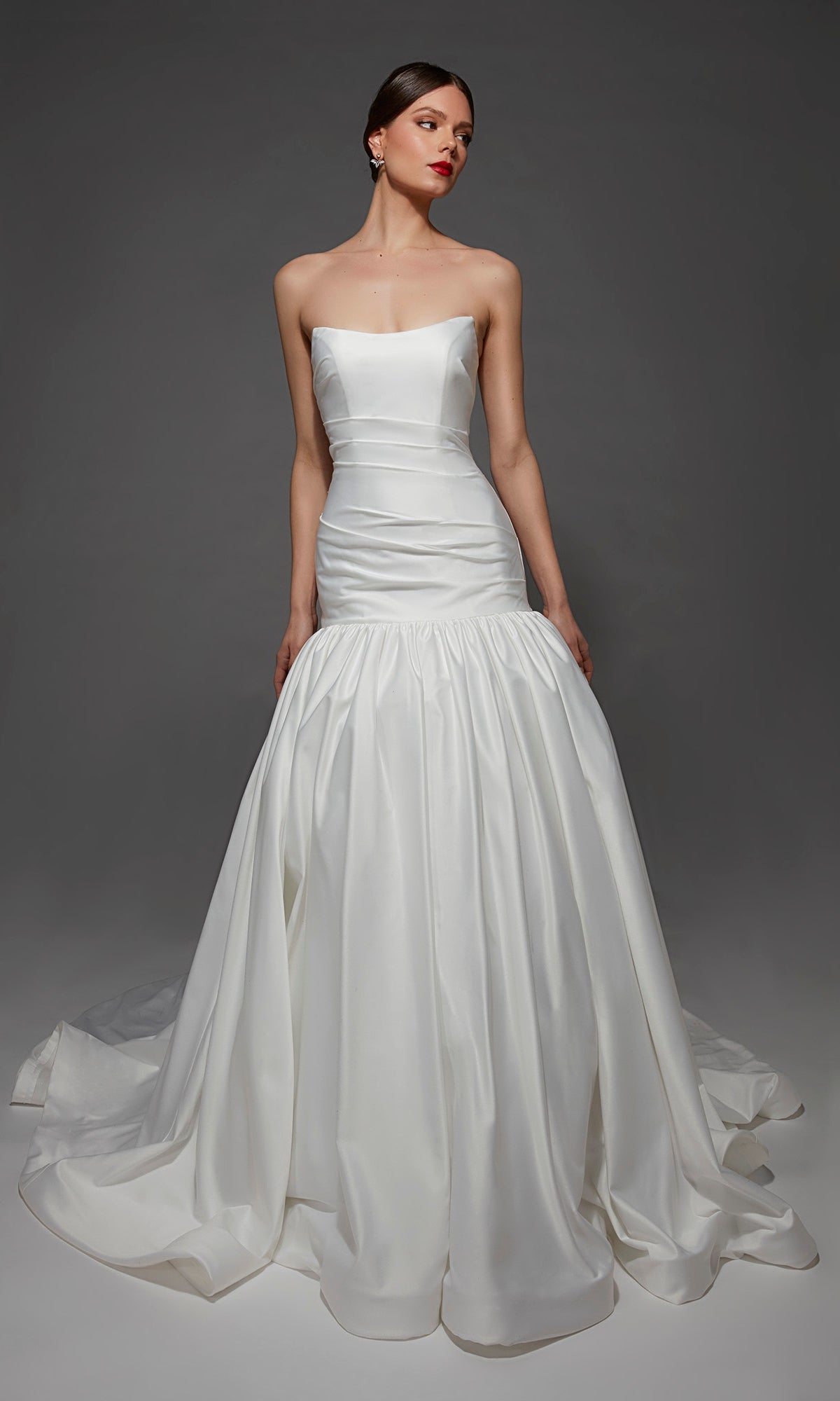 A woman stands against a plain gray background wearing the Alyce 7181 long wedding dress, which features a strapless satin design with a fitted bodice and a full, flared mermaid skirt.