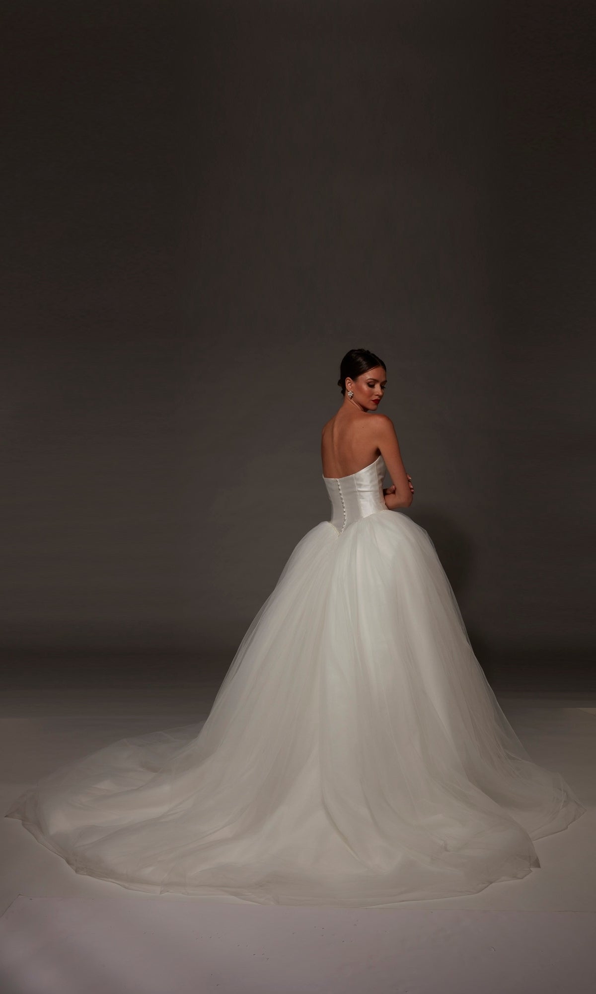A woman stands facing away, looking over her shoulder, against a dark background, wearing the Alyce 7182 long wedding dress—a strapless white ball gown with a mikado bodice.