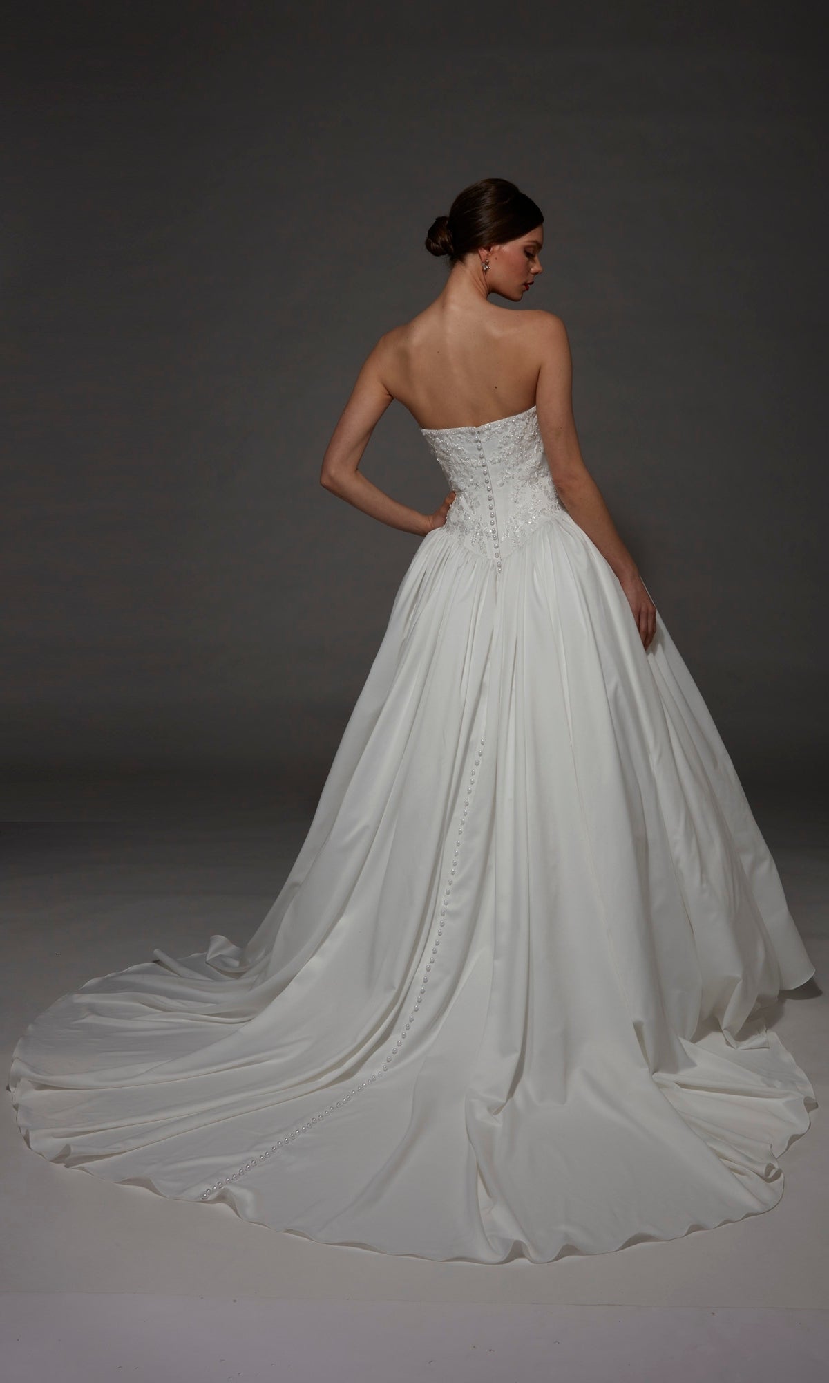 A woman wearing the Alyce 7188 long wedding dress—a strapless satin ball gown with a sweetheart neckline and a sweeping train—stands against a dark background, facing away from the camera.