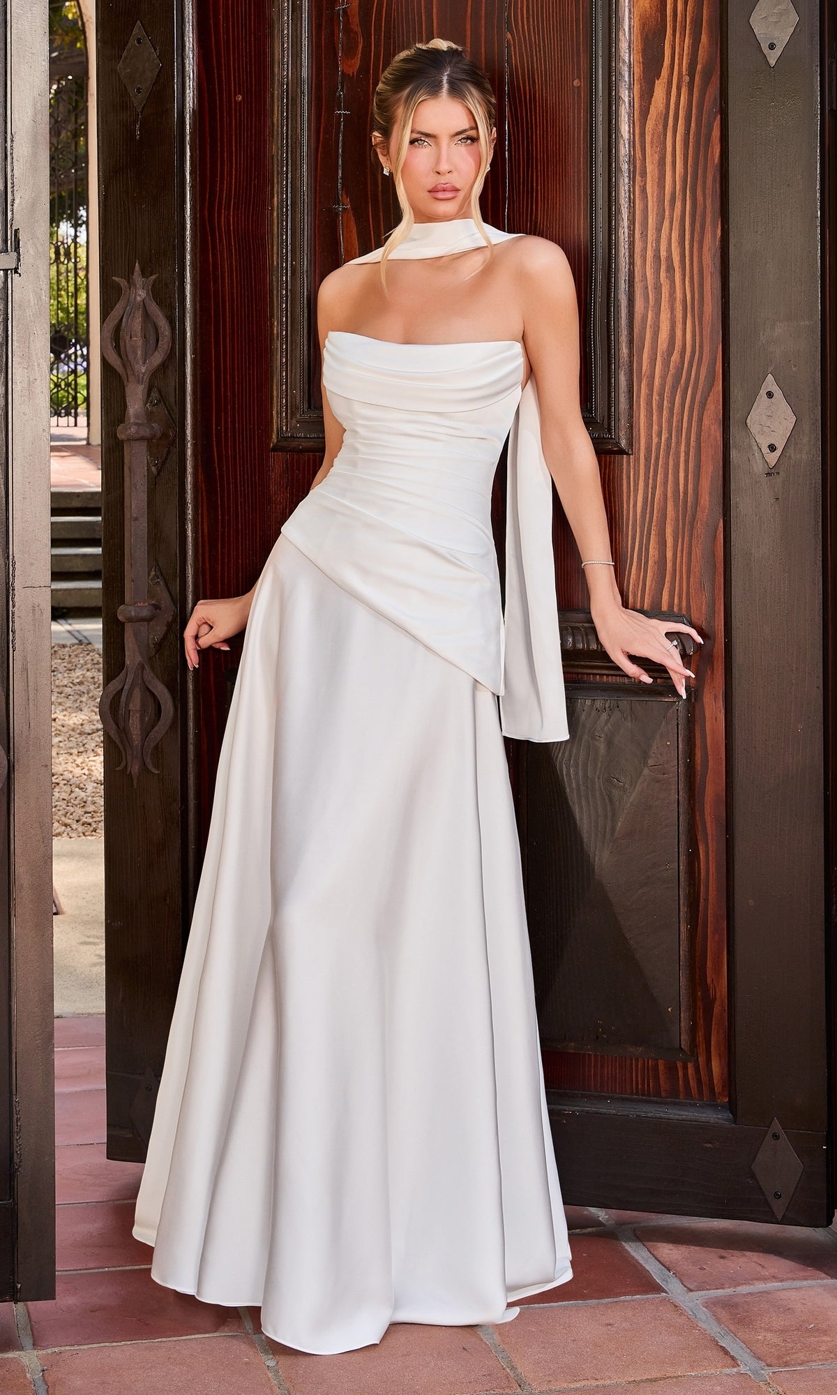A woman in the Ladivine 7432W Long Prom Dress in Off White, featuring a stretch satin bodice and strapless silhouette, stands before a large wooden doorway, facing forward with one hand resting on the door frame.