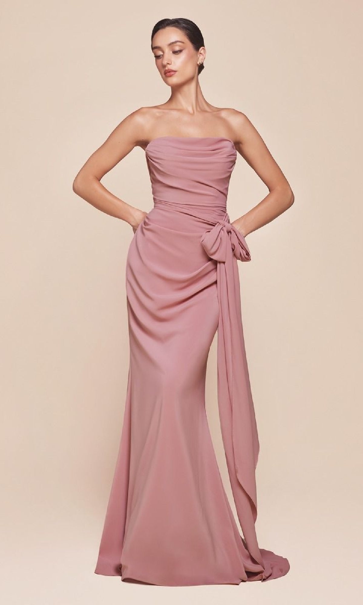A woman stands against a beige background wearing the Ladivine 7433 Multi-Way Long Chiffon Prom Dress, featuring a strapless mauve design with a draped bodice and large side bow detail.