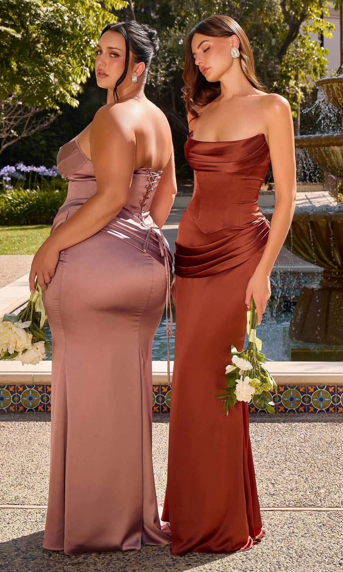 Two women stand side by side outdoors near a fountain, each holding white flowers, wearing the Ladivine 7439 long prom dress. One wears it in mauve with a boned corset top, while the other wears it in rust with a delicate lace-up back.