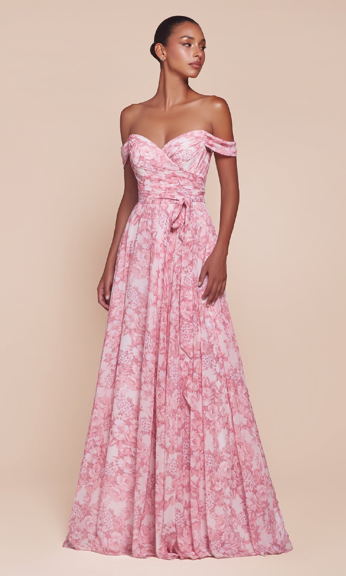 A woman stands against a neutral background wearing the Long Floral-Print A-Line Prom Dress: Ladivine 7441, a floor-length pink gown with off-the-shoulder sleeves, a sweetheart neckline, floral print, and a tied sash at the waist.