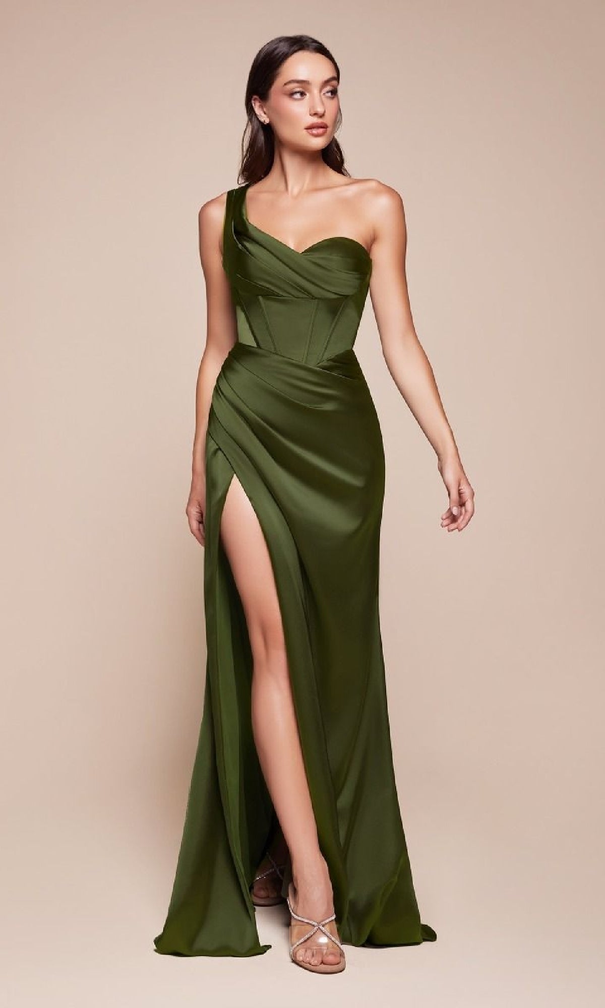A woman in the Ladivine 7448 One-Shoulder Lace-Up Prom Dress in olive green, featuring a corset bodice and thigh-high slit, pairs it with strappy silver heels while standing against a plain beige background.