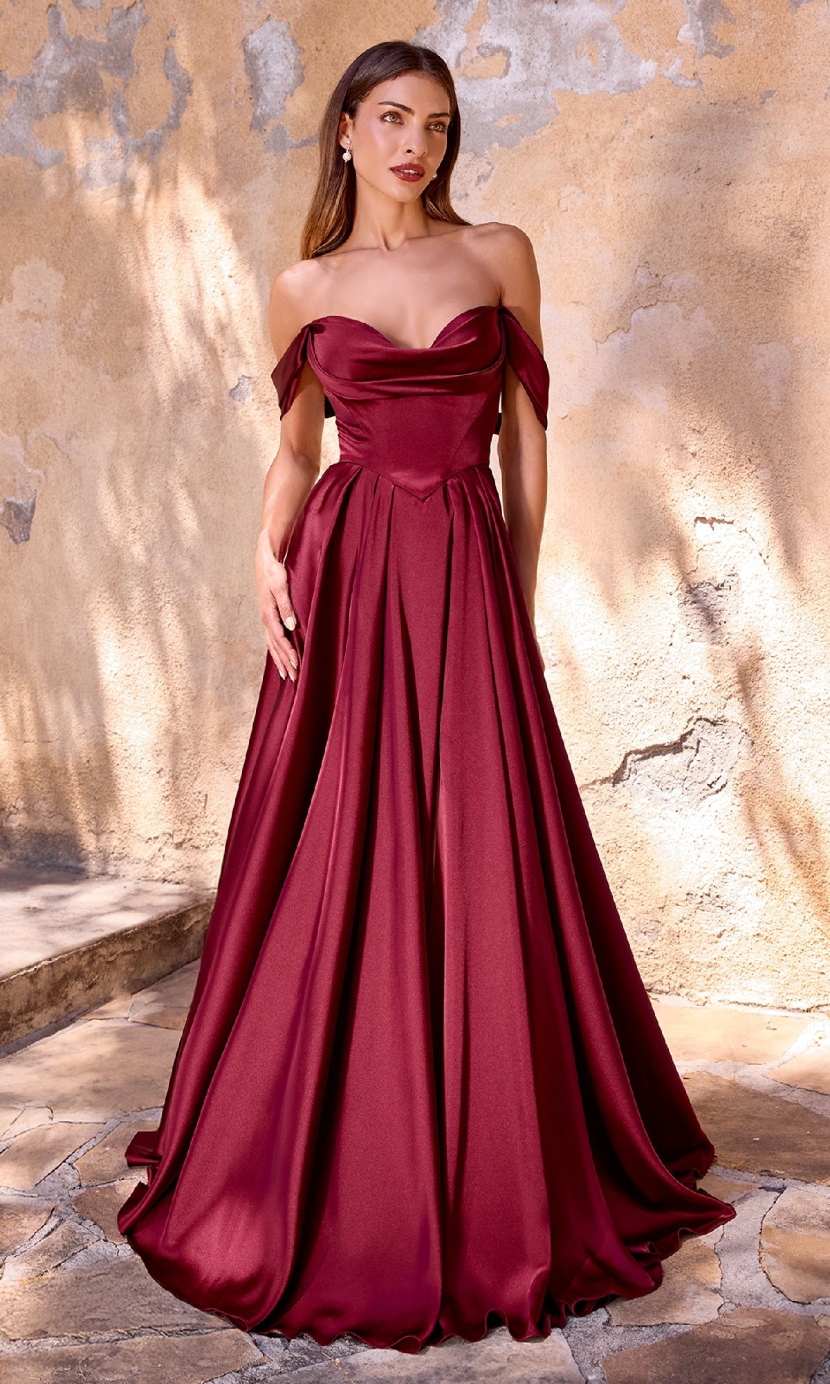 A woman stands before a textured beige wall, wearing the Ladivine 7449 Lace-Up Long Satin Prom Dress—an off-the-shoulder, floor-length burgundy gown with a full skirt.