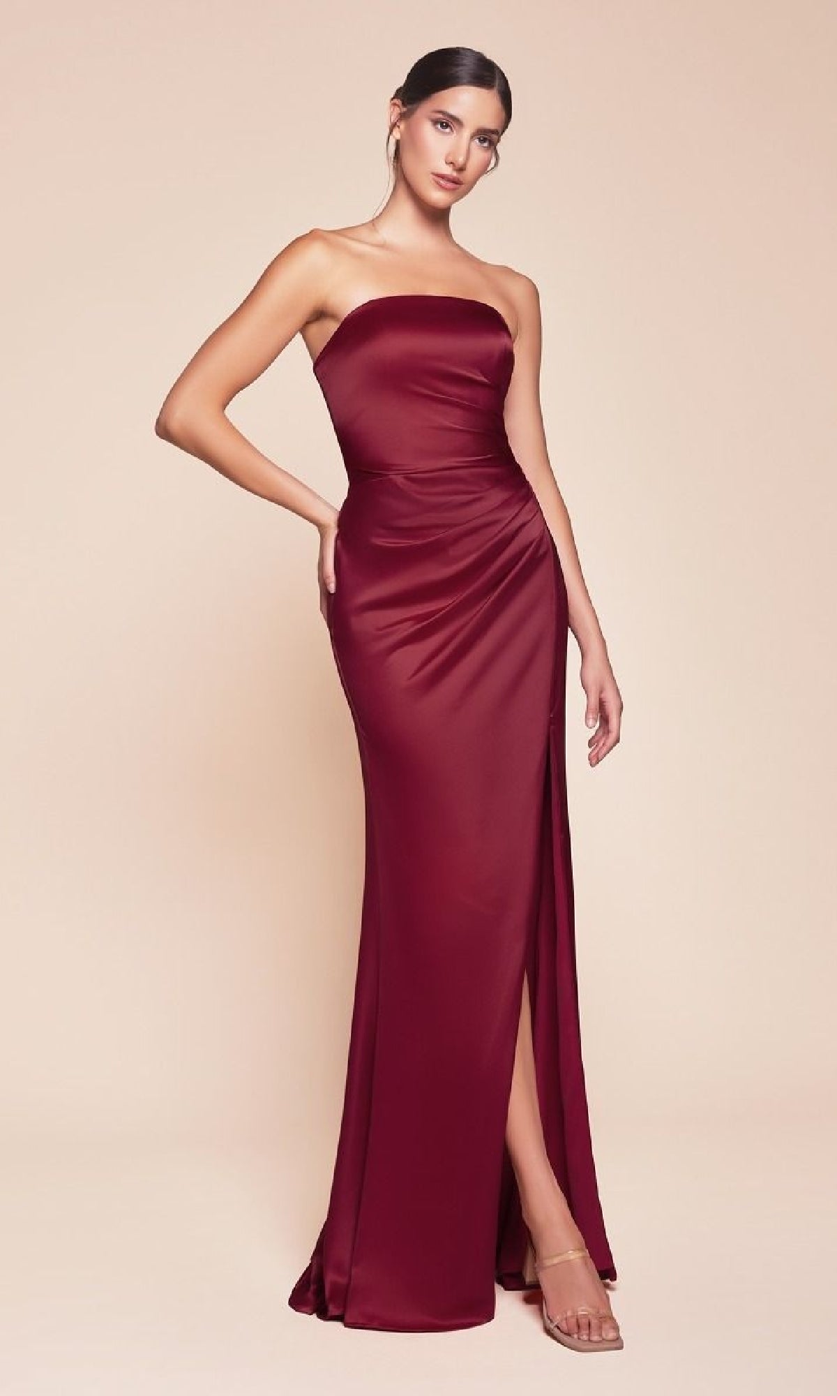 A woman wears the Ladivine 7451, a simple strapless long prom dress in burgundy with a thigh-high slit, standing against a plain beige background.